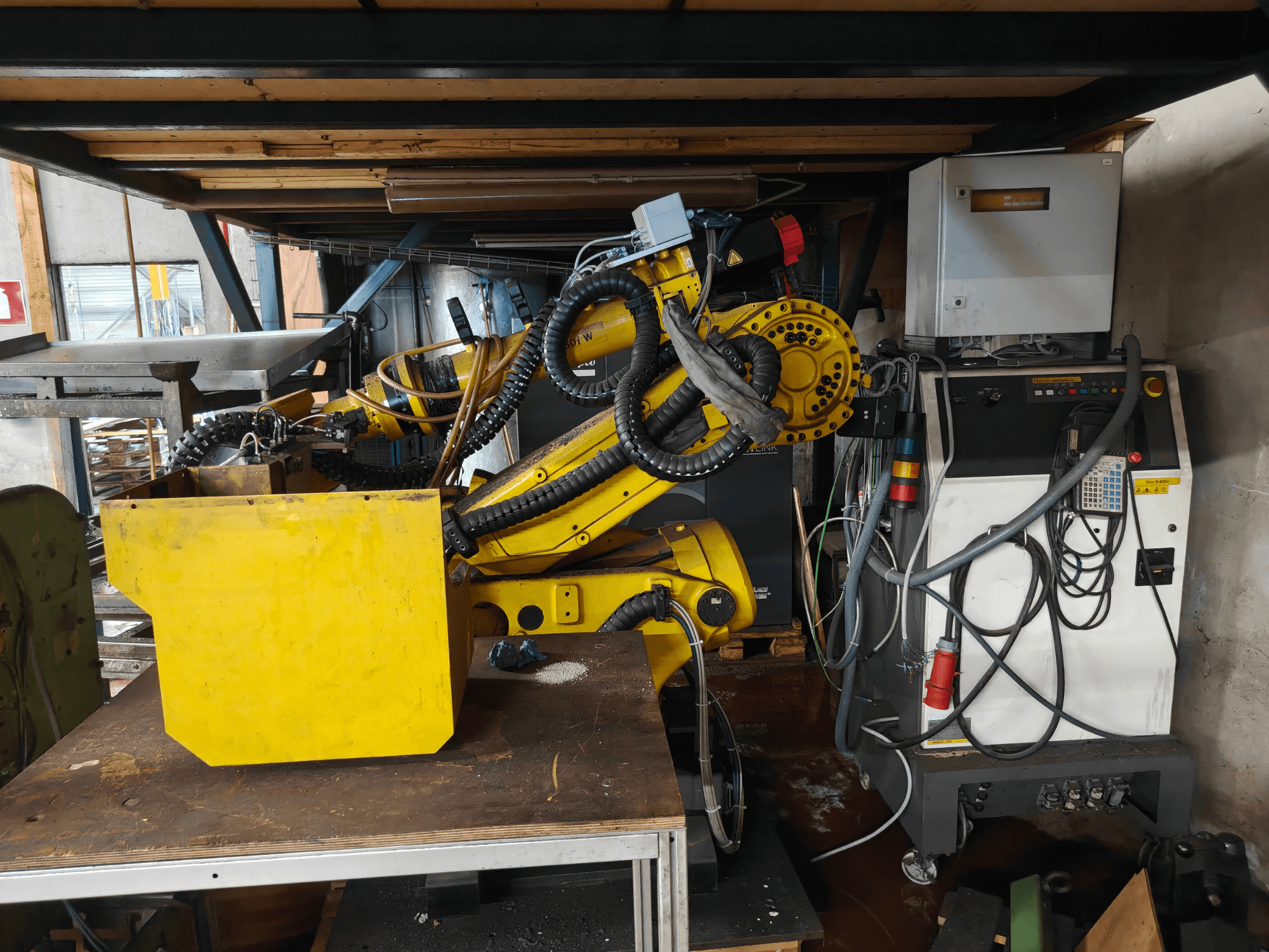 Vista frontal do braço de um robô FANUC S-430i W, de cor amarela, com o painel de controlo, cabos e um componente amarelo sobre uma bancada de trabalho.