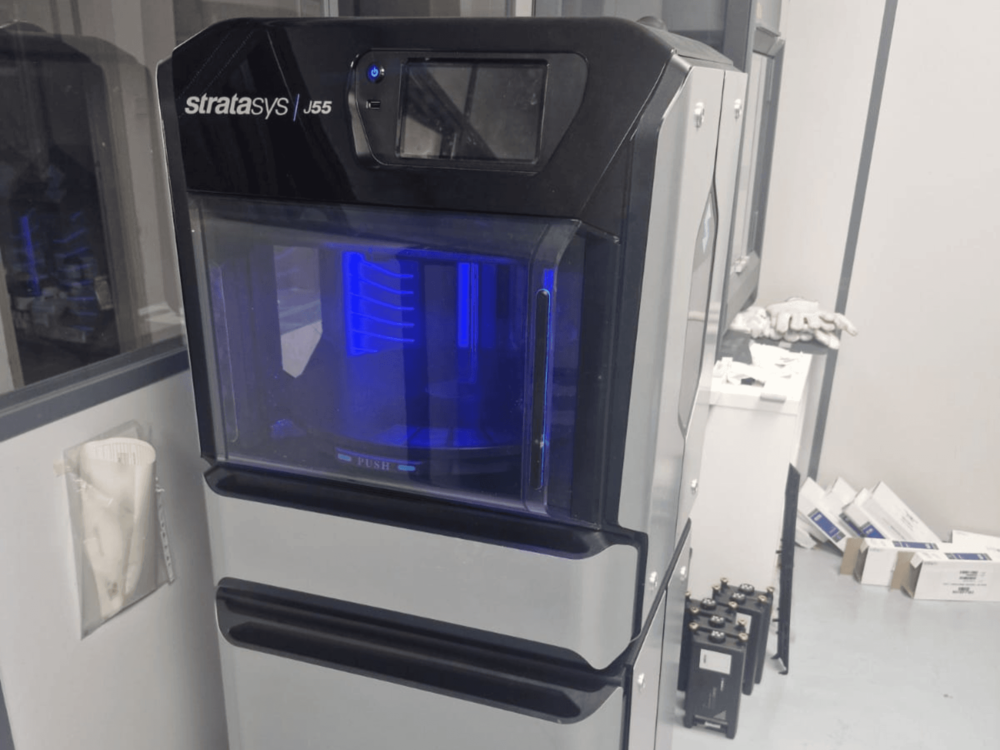 Impressora 3D de plástico Stratasys J55 Prime numa vista lateral, mostrando o seu design elegante e a área de impressão iluminada.