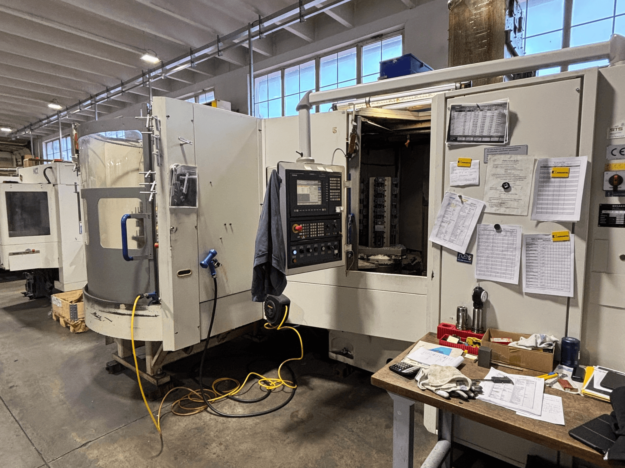 Máquina CNC numa oficina, vista frontal, com painel de controlo e documentação ao lado; ferramentas e materiais sobre a mesa.