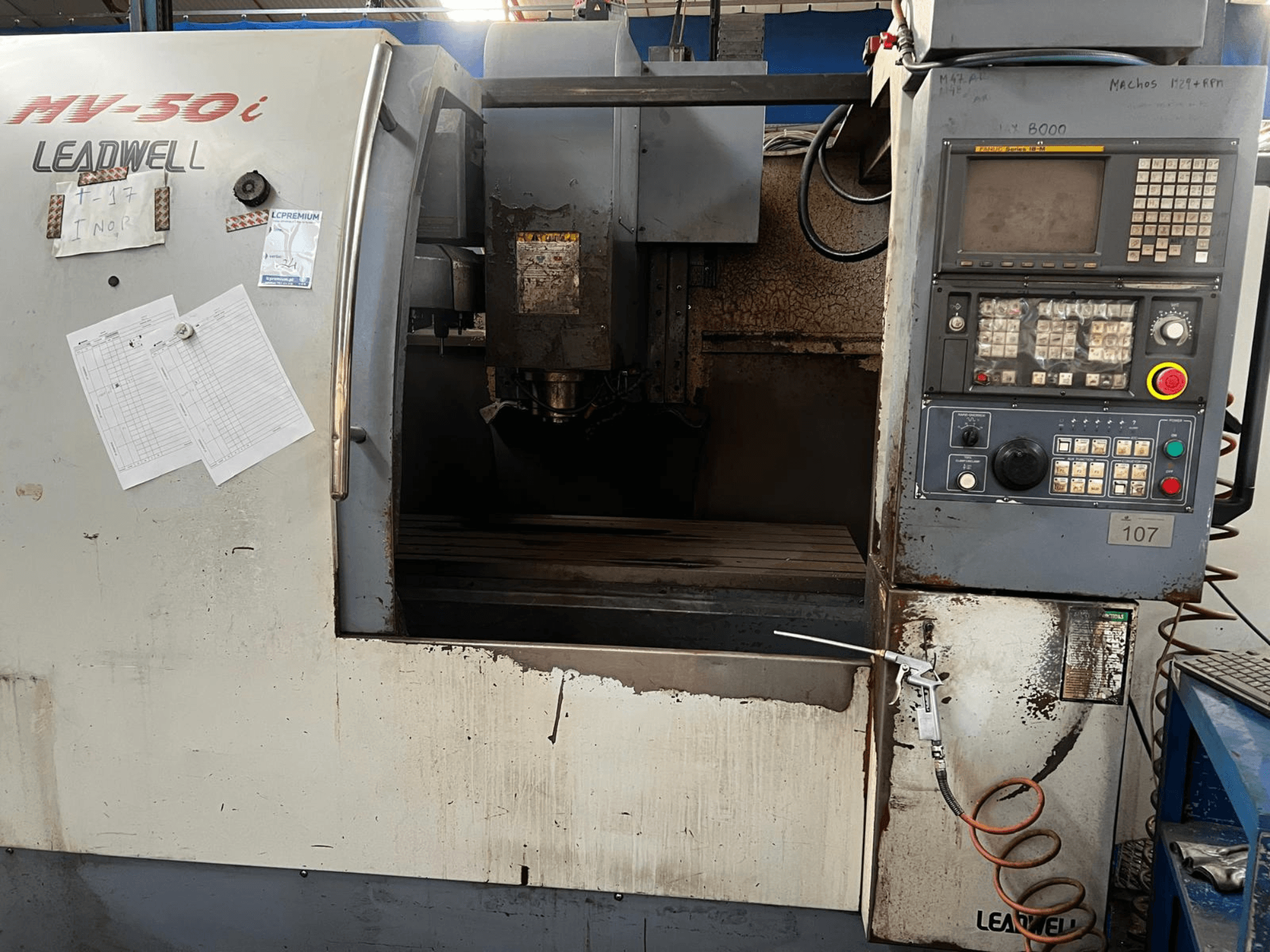 Centro de maquinagem vertical Leadwell MV-50i, vista frontal, mostrando o painel de controlo, a área do fuso e a mesa de trabalho com sinais de desgaste.