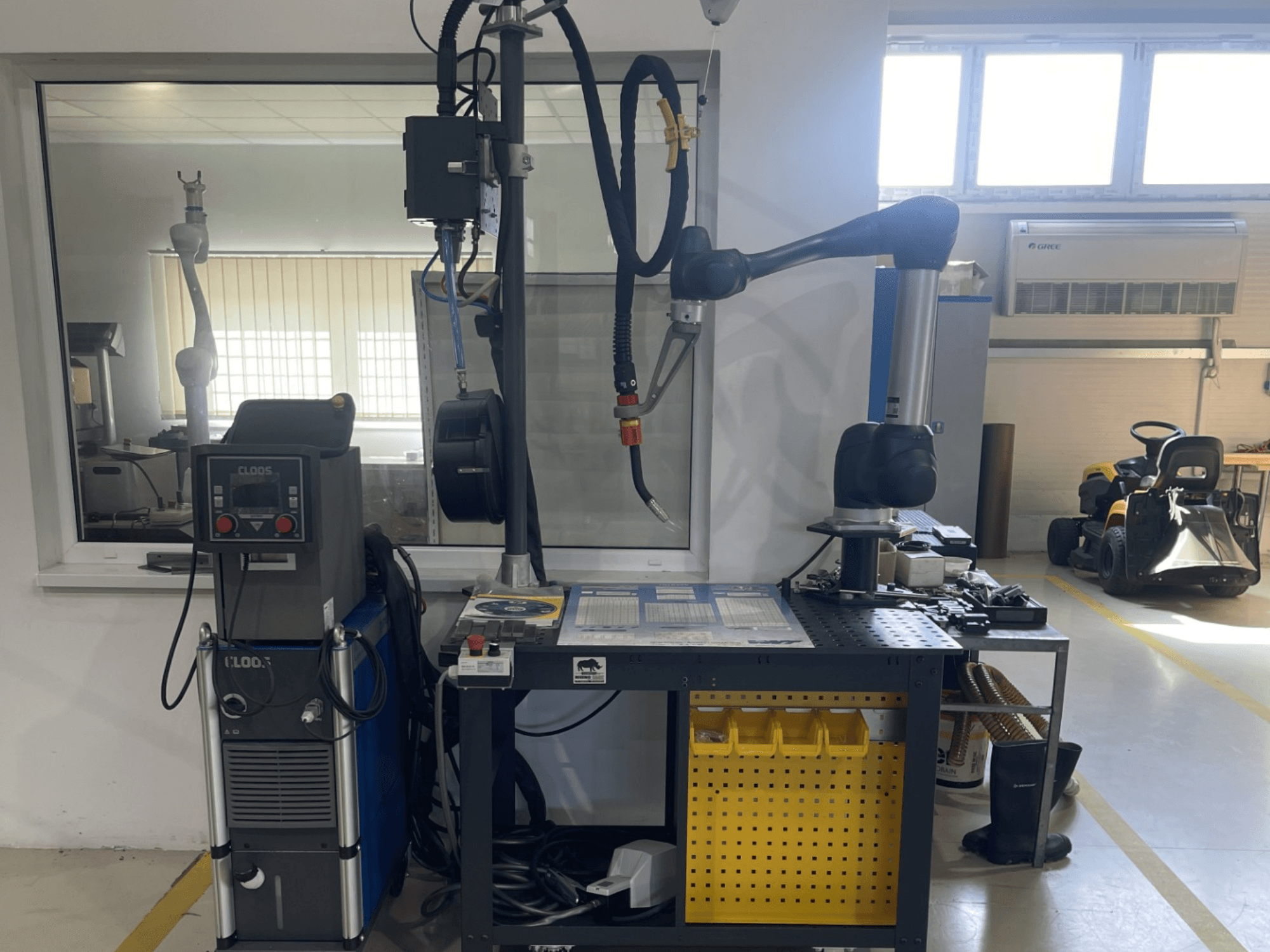 Robô colaborativo DOOSAN M1013 numa oficina, vista lateral com equipamento de soldadura e ferramentas numa bancada de trabalho.