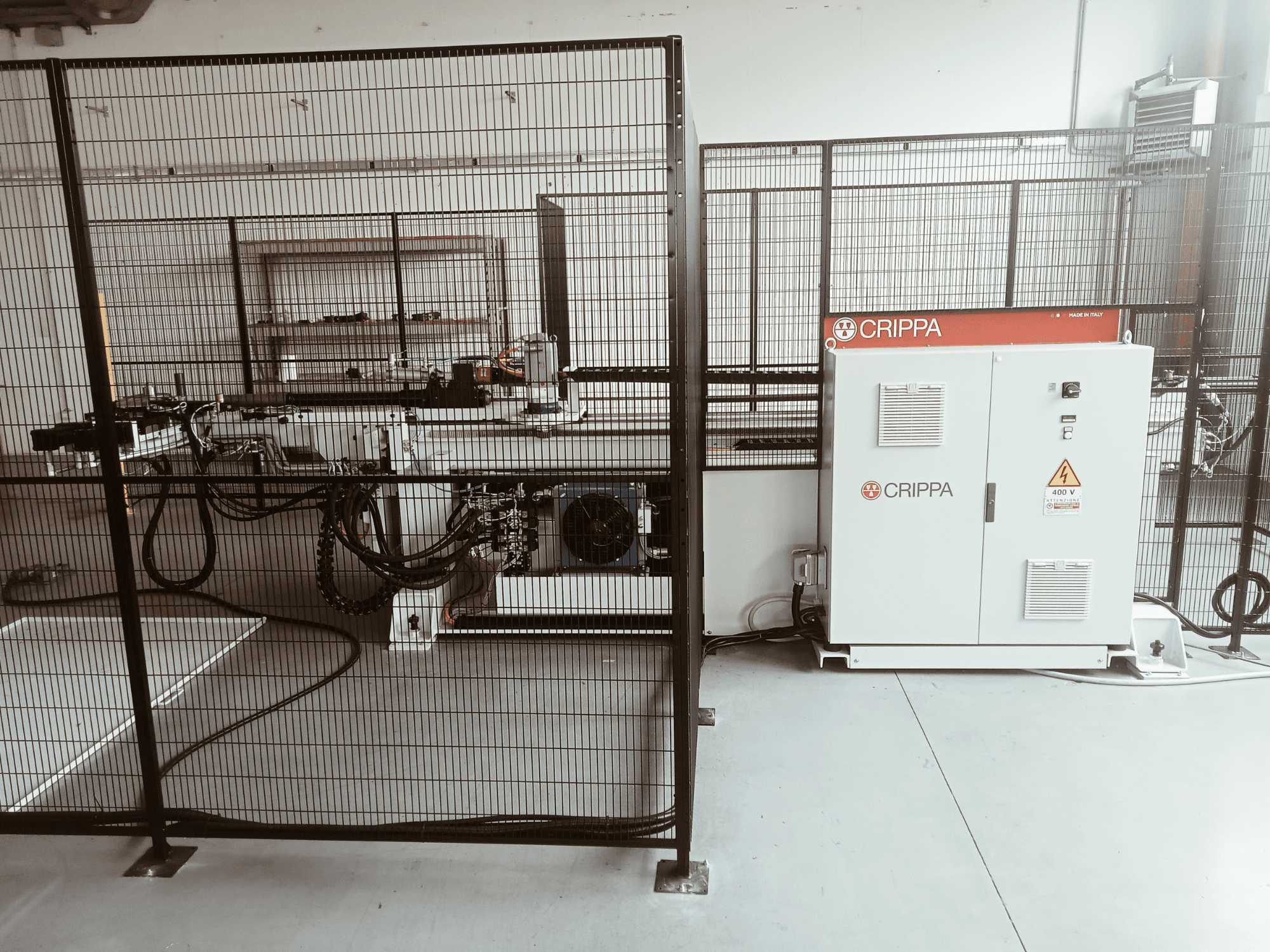 Máquina CRIPPA branca atrás de uma gaiola de segurança, com mangueiras e componentes visíveis, vista lateral, ambiente industrial.