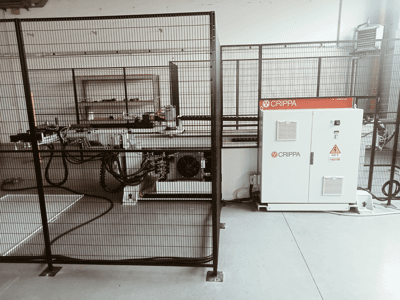 Máquina CRIPPA branca atrás de uma gaiola de segurança, com mangueiras e componentes visíveis, vista lateral, ambiente industrial.