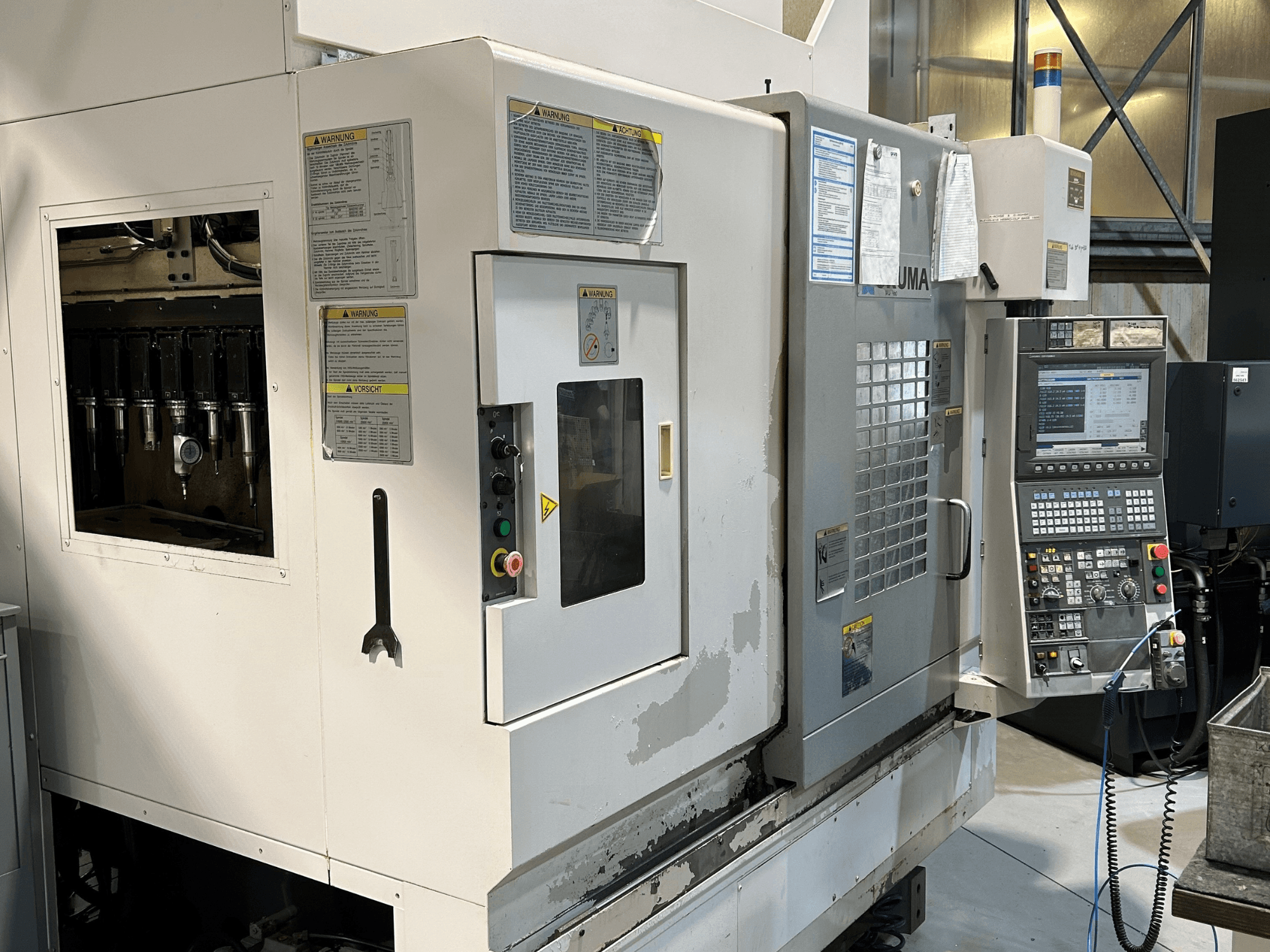 Vista frontal de uma máquina CNC UMA com suportes de ferramentas visíveis, painel de controlo, etiquetas de aviso e botões operacionais.