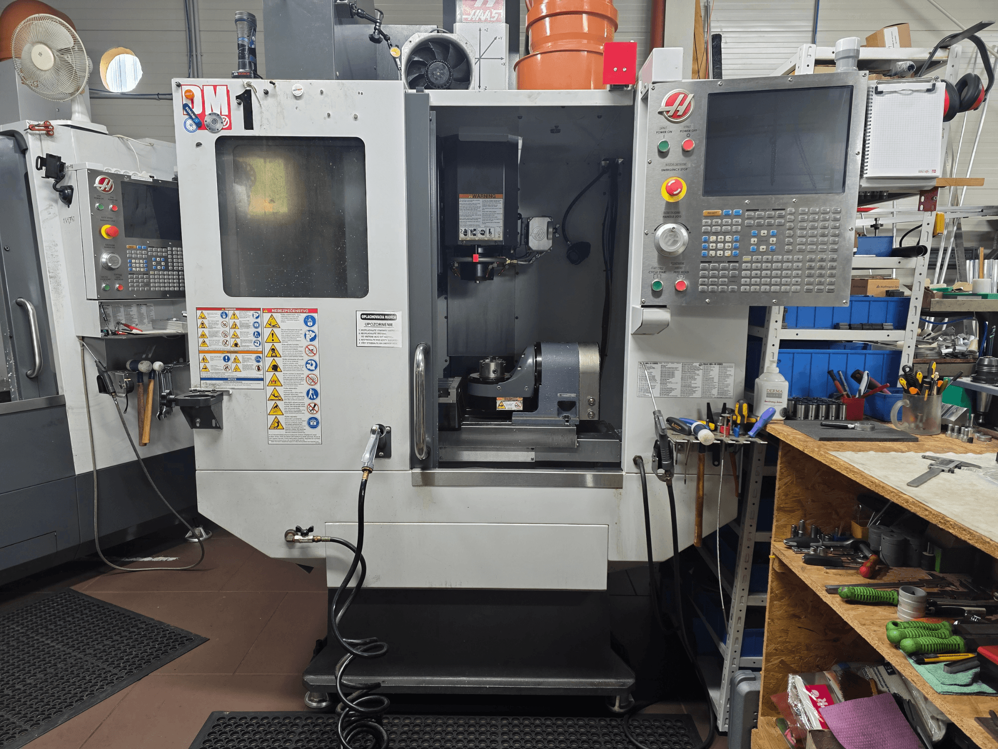 Máquina CNC Haas modelo VF-2, vista frontal, mostrando controlos, área do spindle, etiquetas de segurança e ferramentas numa bancada de trabalho próxima.