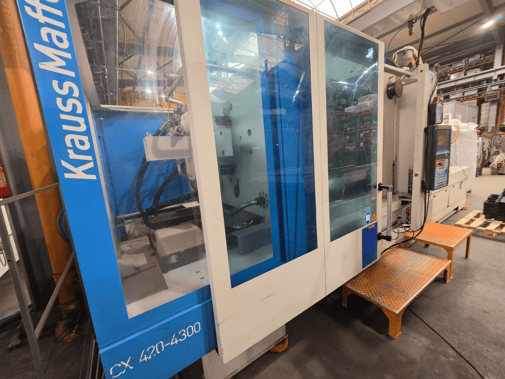 Máquina de moldagem por injeção hidráulica Krauss Maffei CX 420-4300, vista lateral, mostrando o painel de controlo e os painéis transparentes.