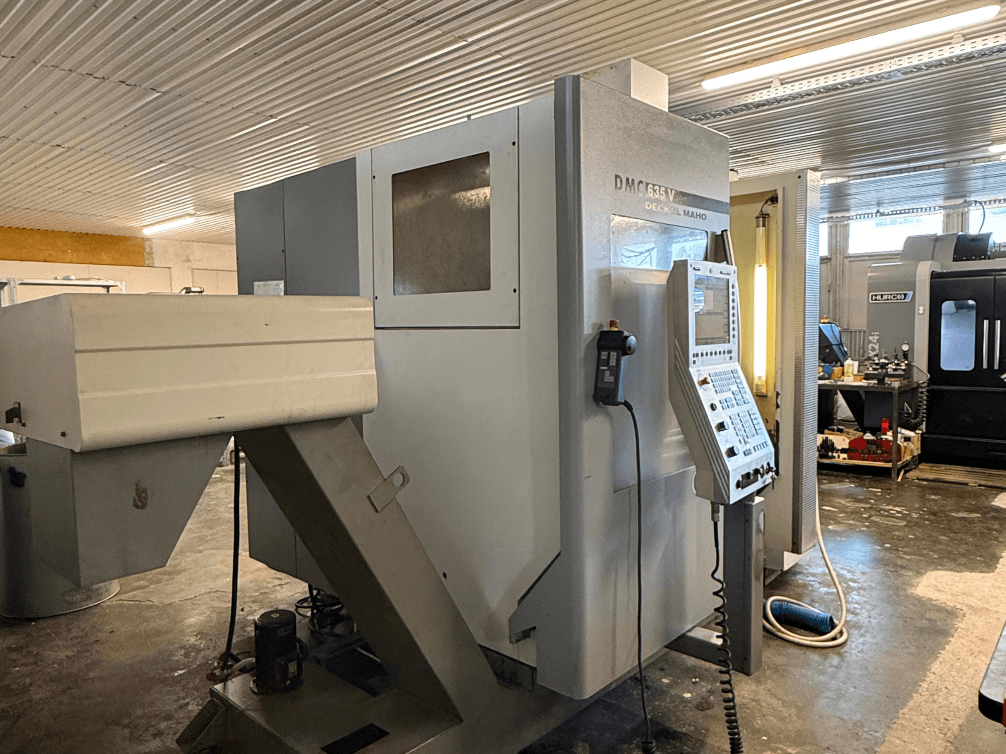 Fresadora DMC G635V numa oficina, vista frontal com painel de controlo e ferramentas eléctricas ligadas visíveis.