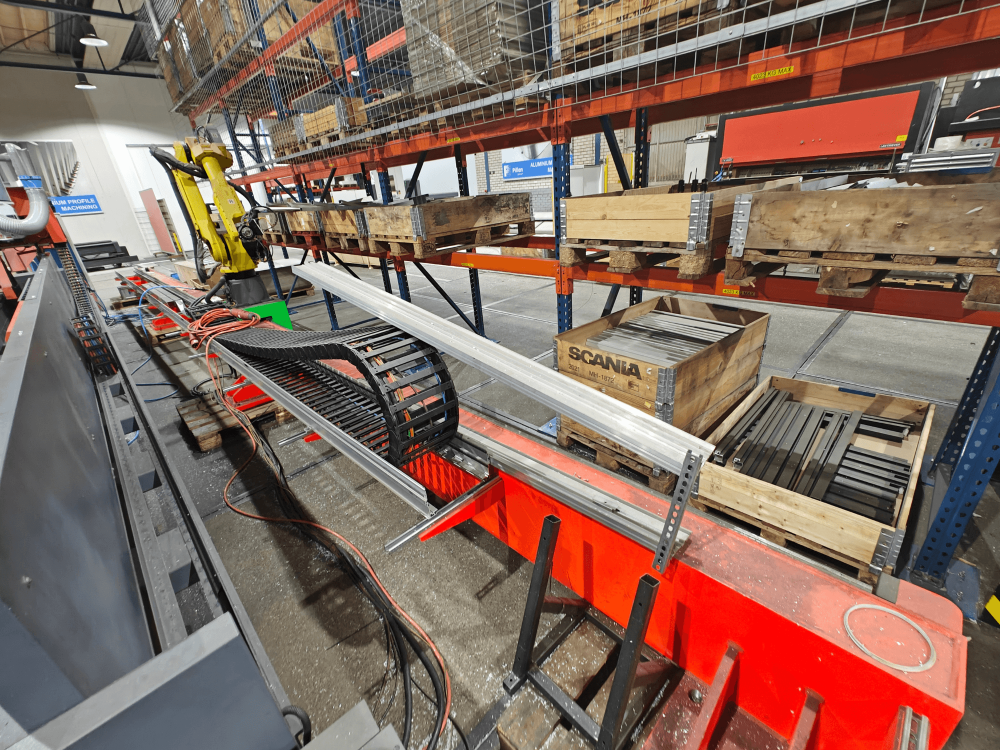 Braço robótico na estação de trabalho, cabos laranja e pretos visíveis, com caixas Scania e perfis de alumínio num ambiente de fábrica.