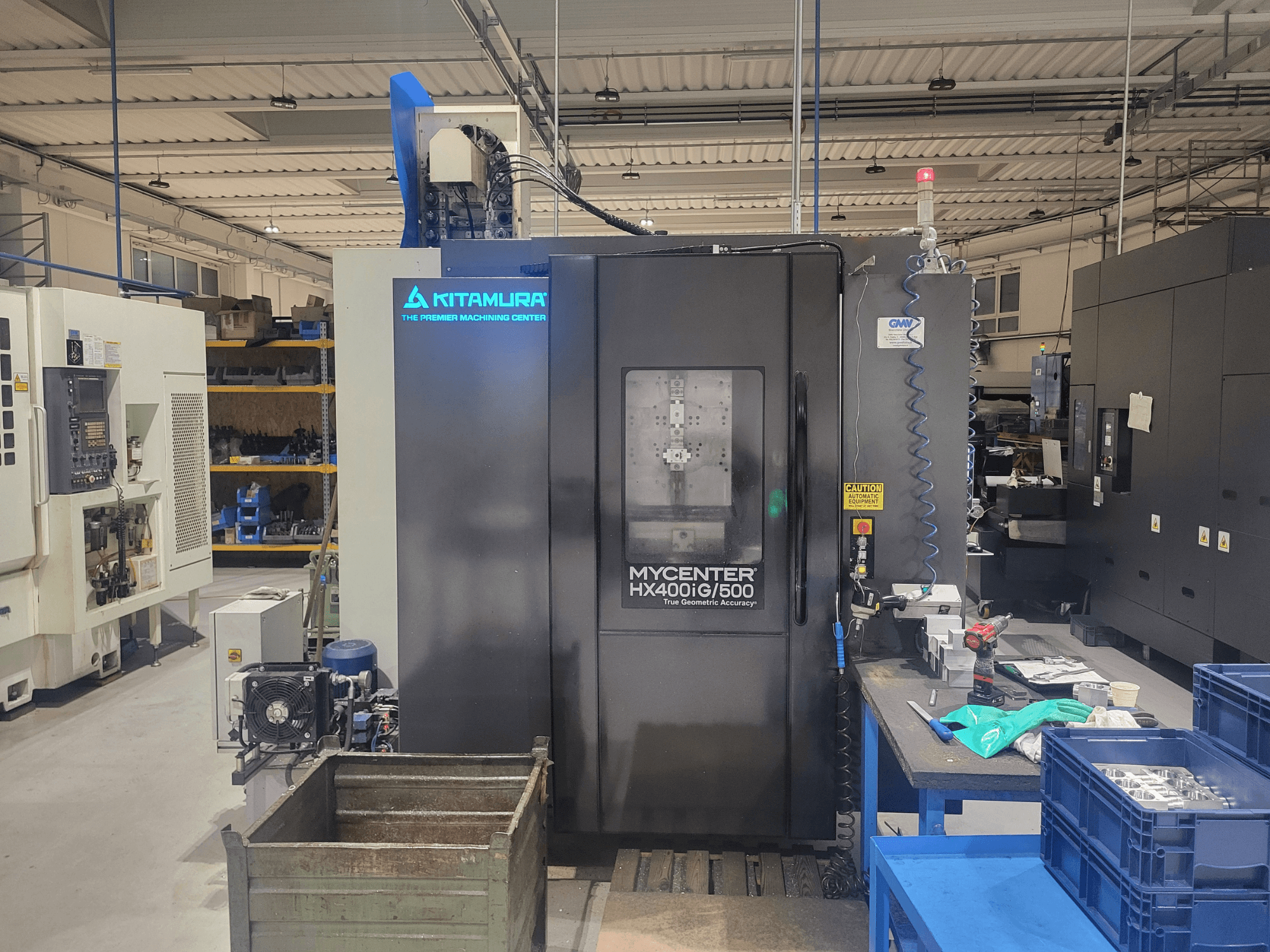Máquina CNC Kitamura Mycenter HX400iG/500 numa oficina, vista frontal, com controlos, armazenamento de ferramentas e materiais nas proximidades.