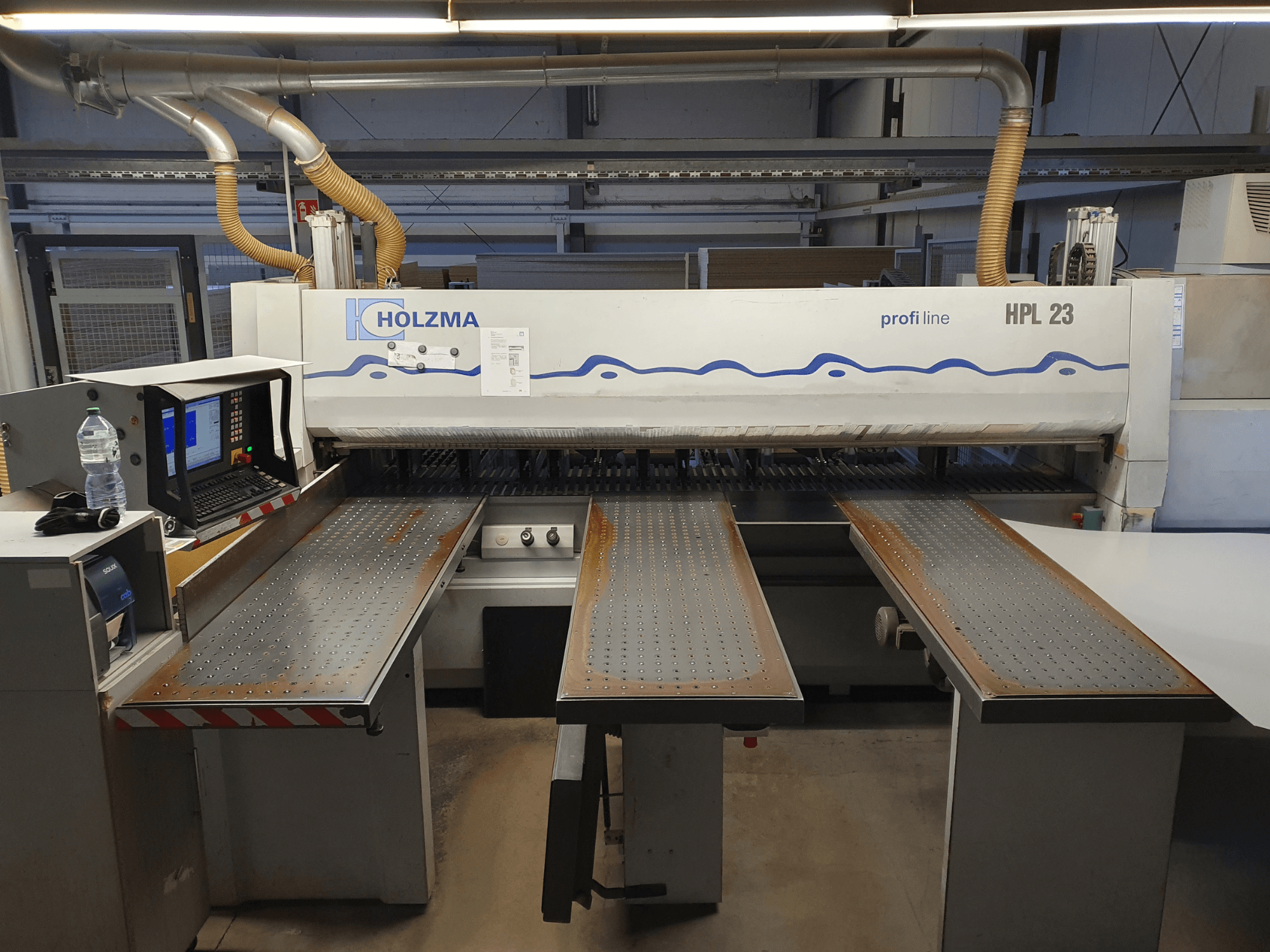 Máquina de corte HOLZMA HPL 23, vista de frente, com três mesas perfuradas, terminal de computador e garrafa de água nas proximidades.