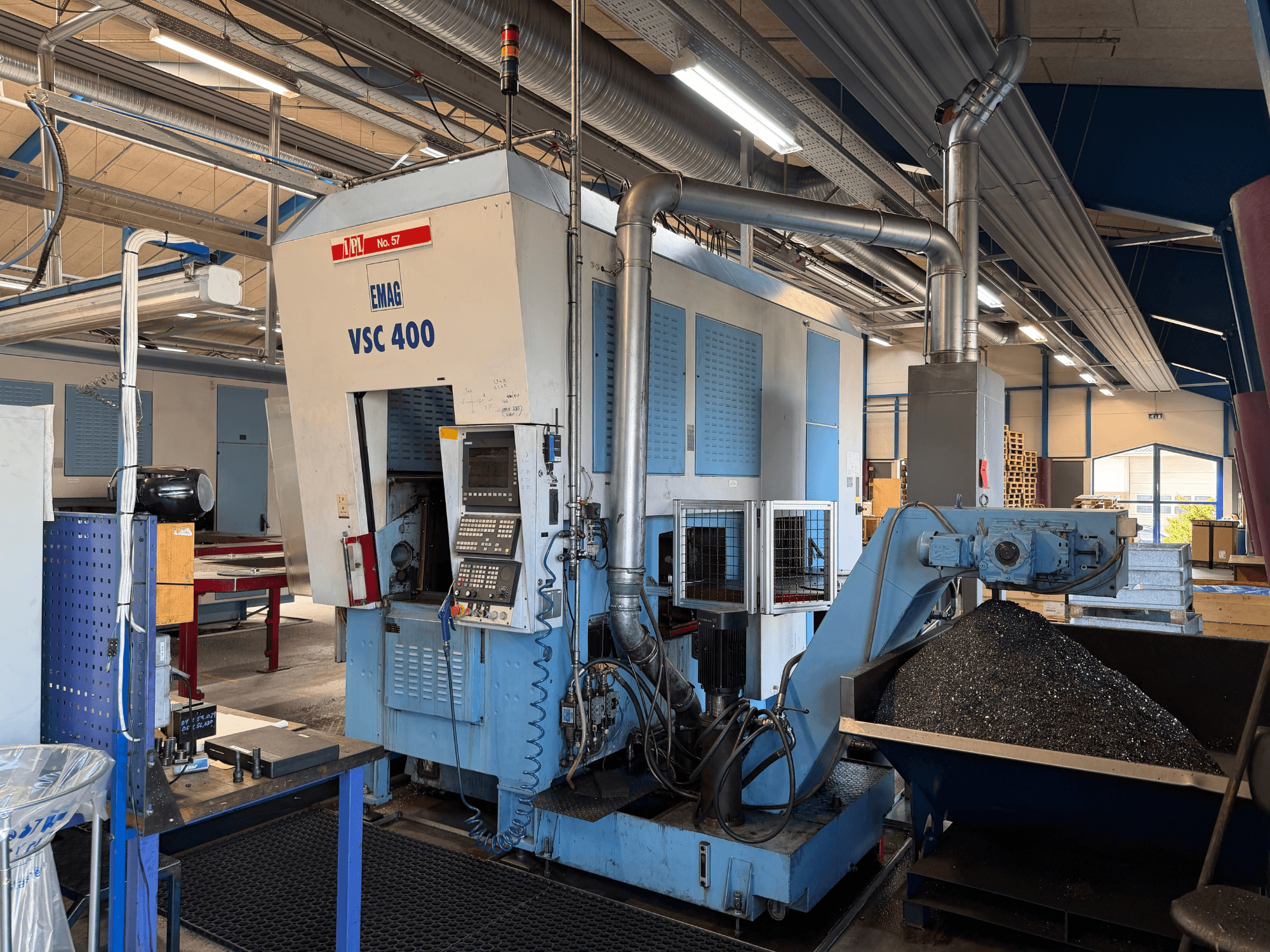 Máquina de torneamento vertical EMAG VSC 400, vista frontal, mostrando o painel de controlo e o transportador de aparas. Ambiente industrial visível.