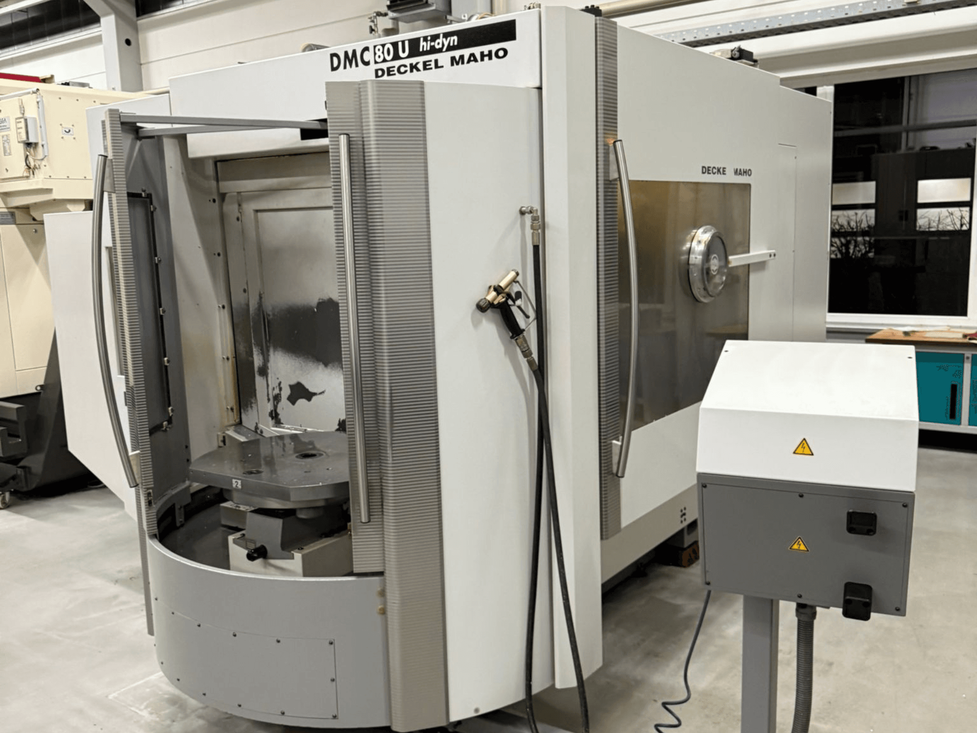 Vista frontal de uma máquina CNC DECKEL MAHO DMC 80 U com portas abertas, mostrando a área de trabalho e o painel de controlo, num ambiente de fábrica.