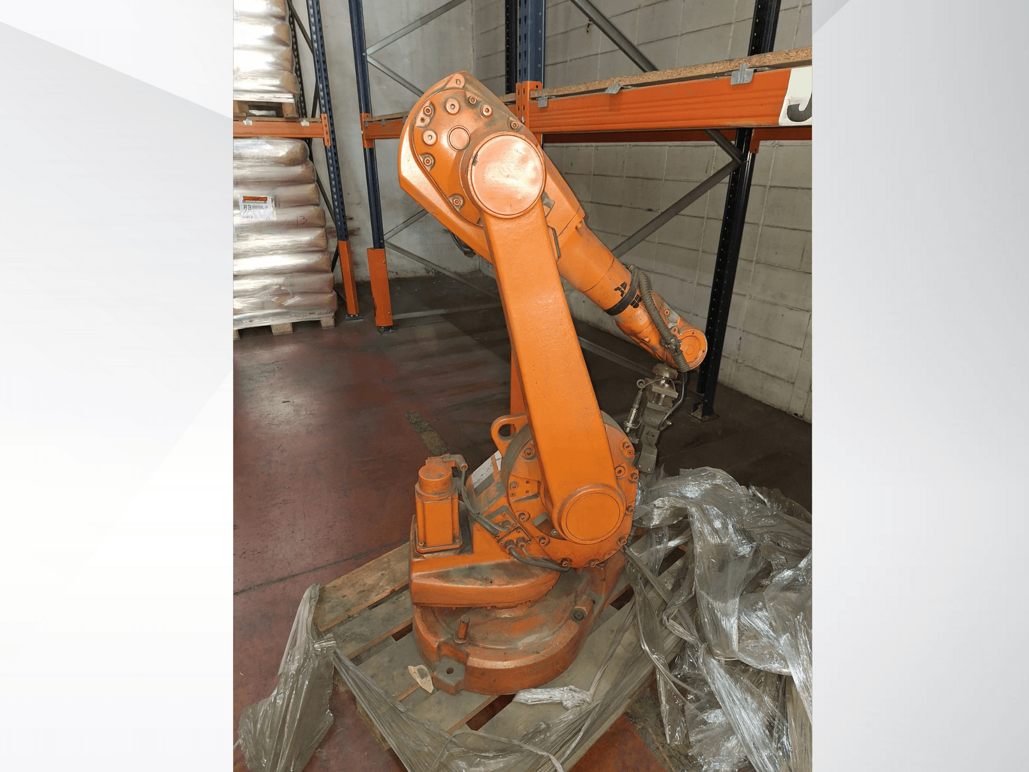 Braço robótico industrial cor de laranja, vista frontal sobre palete de madeira, com cabo ligado e película de plástico de proteção à volta da base.