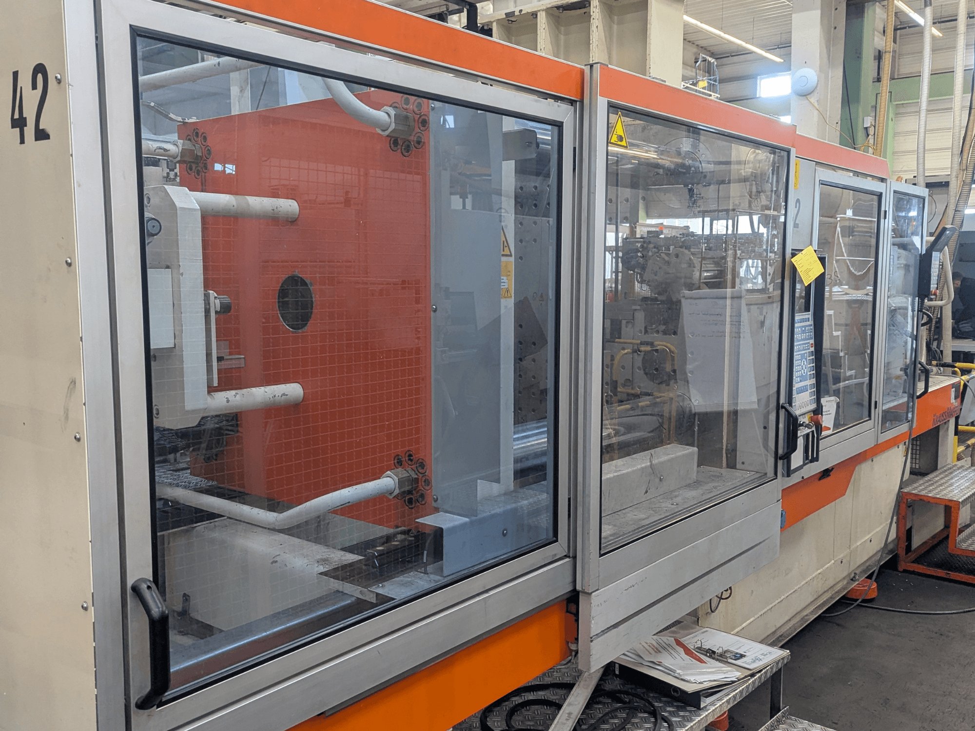 Máquina de moldagem por injeção hidráulica Krauss Maffei KM 350-3000, vista lateral mostrando os controlos e componentes internos atrás de um painel de vidro.