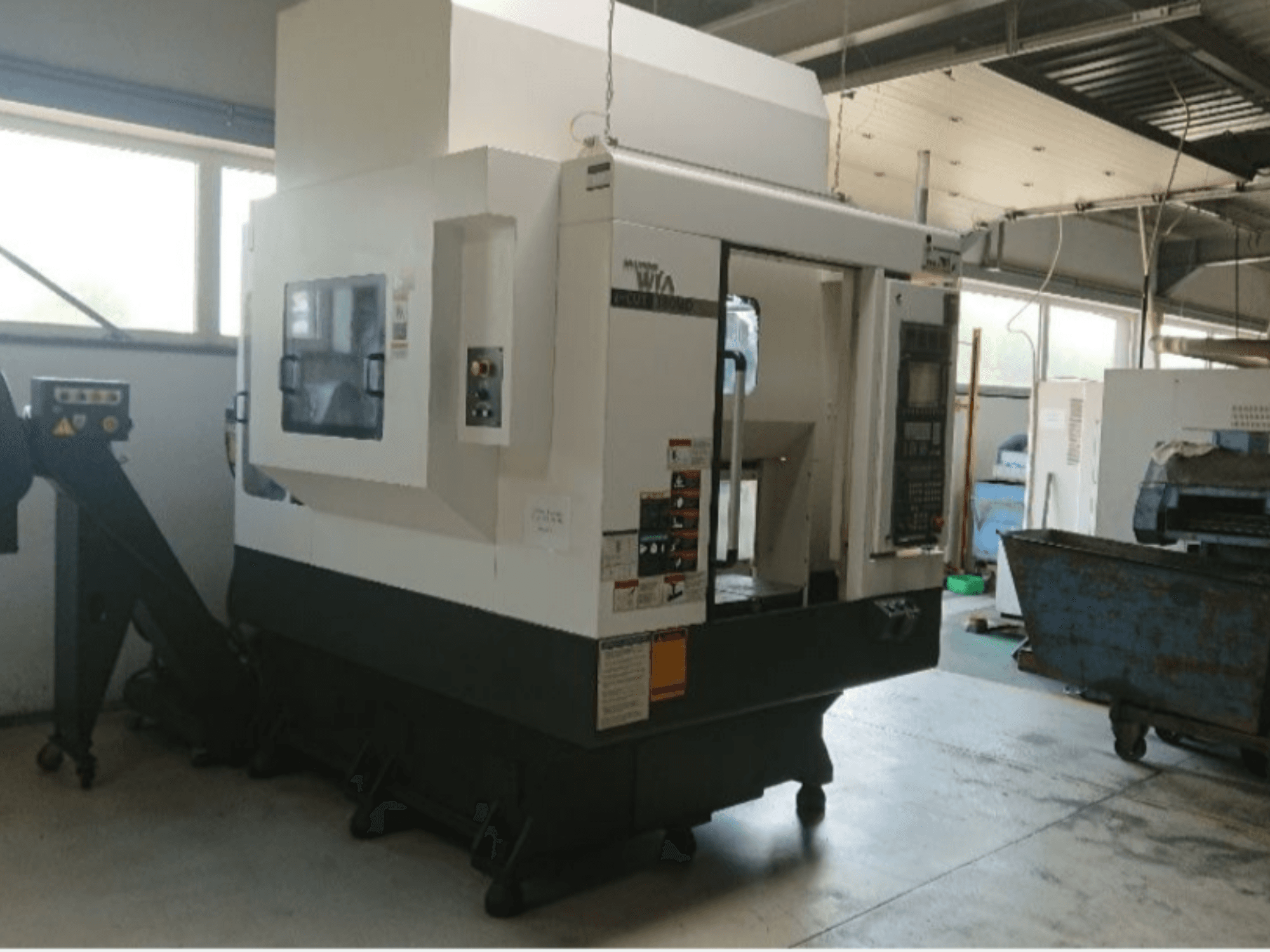 Máquina CNC Haas, vista lateral, com painel de controlo, etiquetas de aviso e uma calha de carga, num ambiente de oficina.
