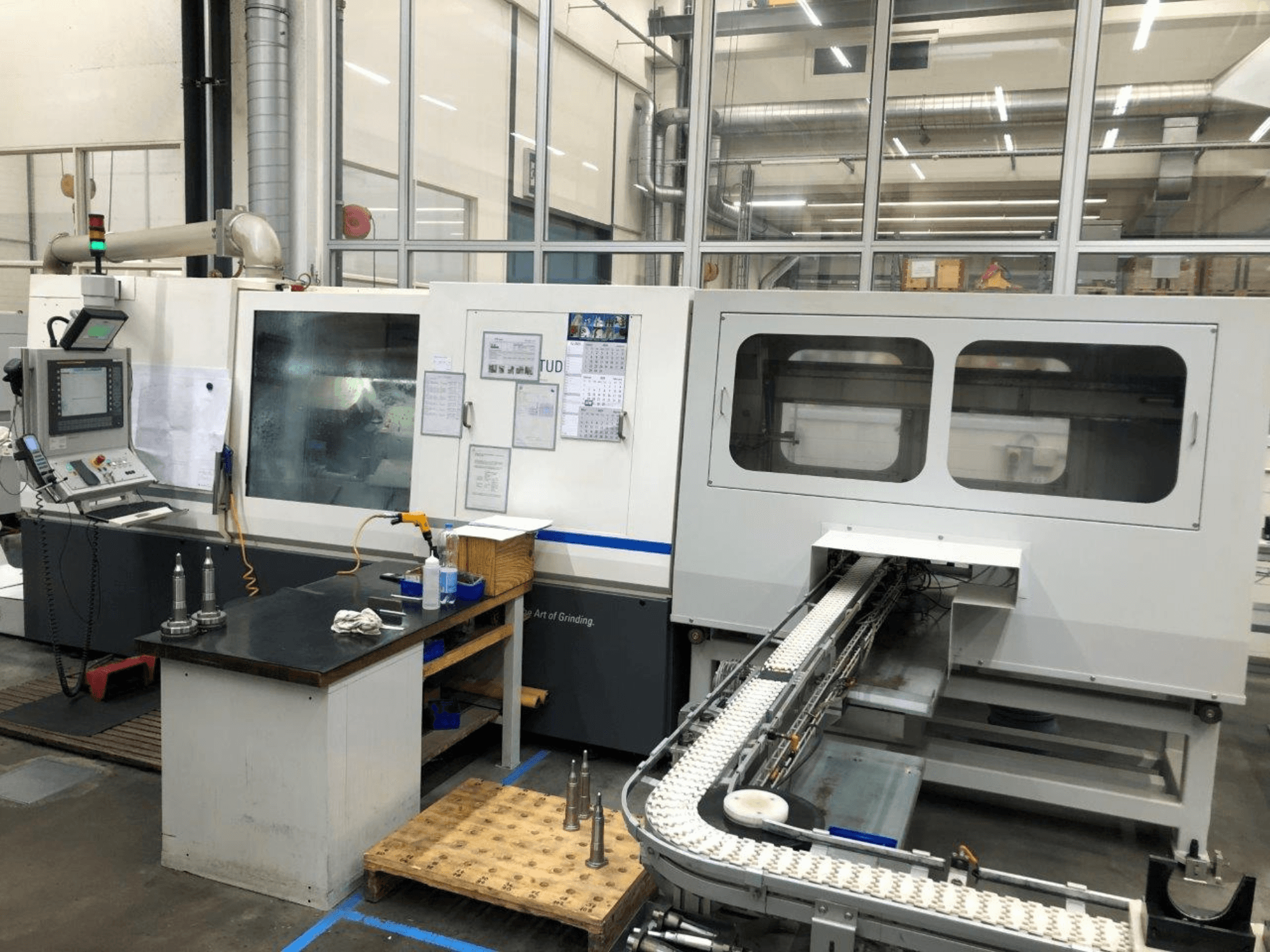 Rectificadora CNC com painel de controlo, vista frontal; possui um tapete rolante e janelas de visualização claras, mostrando ferramentas de precisão.