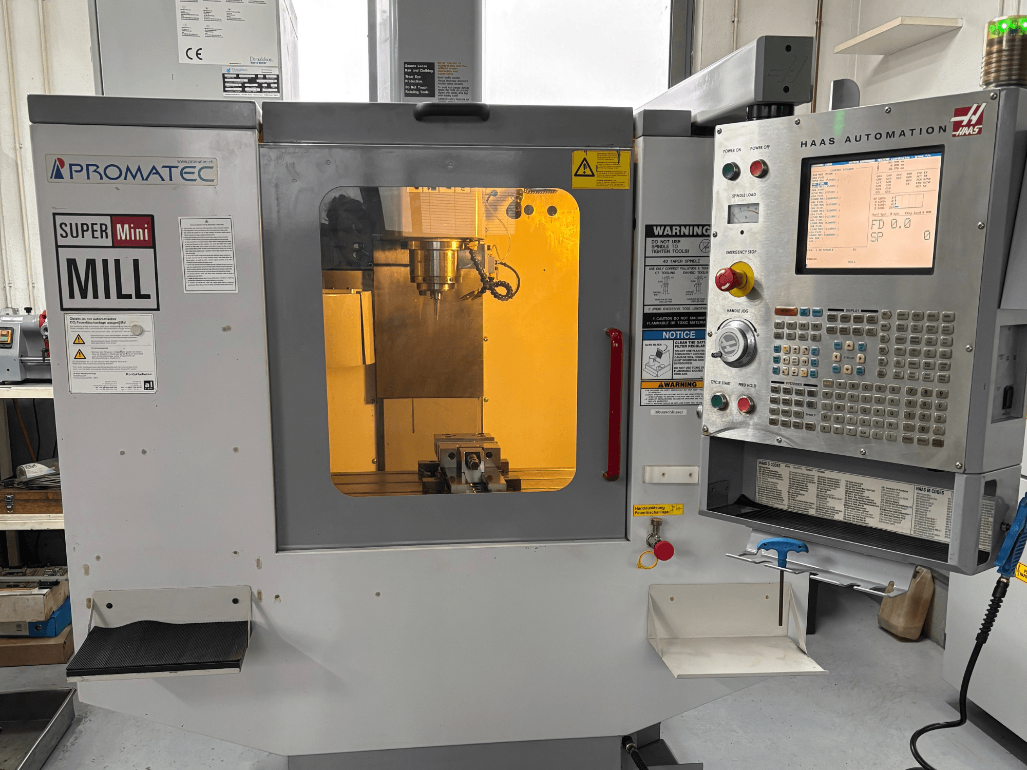Super Mini Fresadora Promatec em vista frontal, equipada com um painel de controlo Haas Automation e uma ferramenta de corte visível no interior.