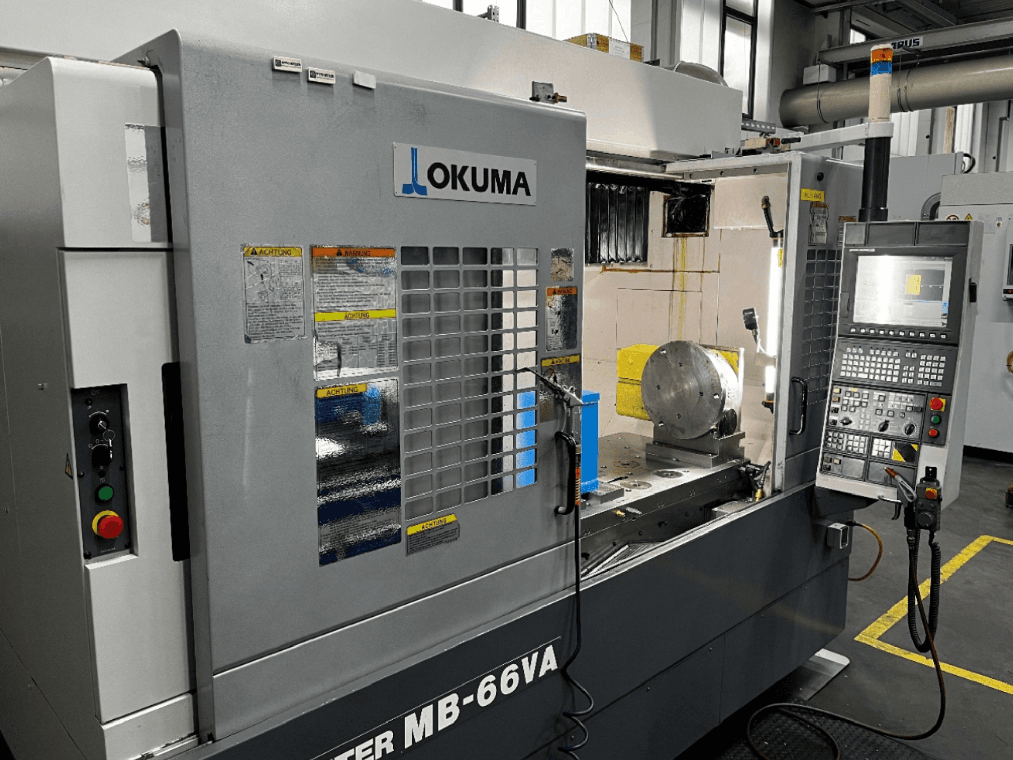 Vista frontal do Centro de Maquinação Vertical Okuma MB66-VA mostrando o painel de controlo, a peça de trabalho e o interior da máquina com etiquetas de segurança.