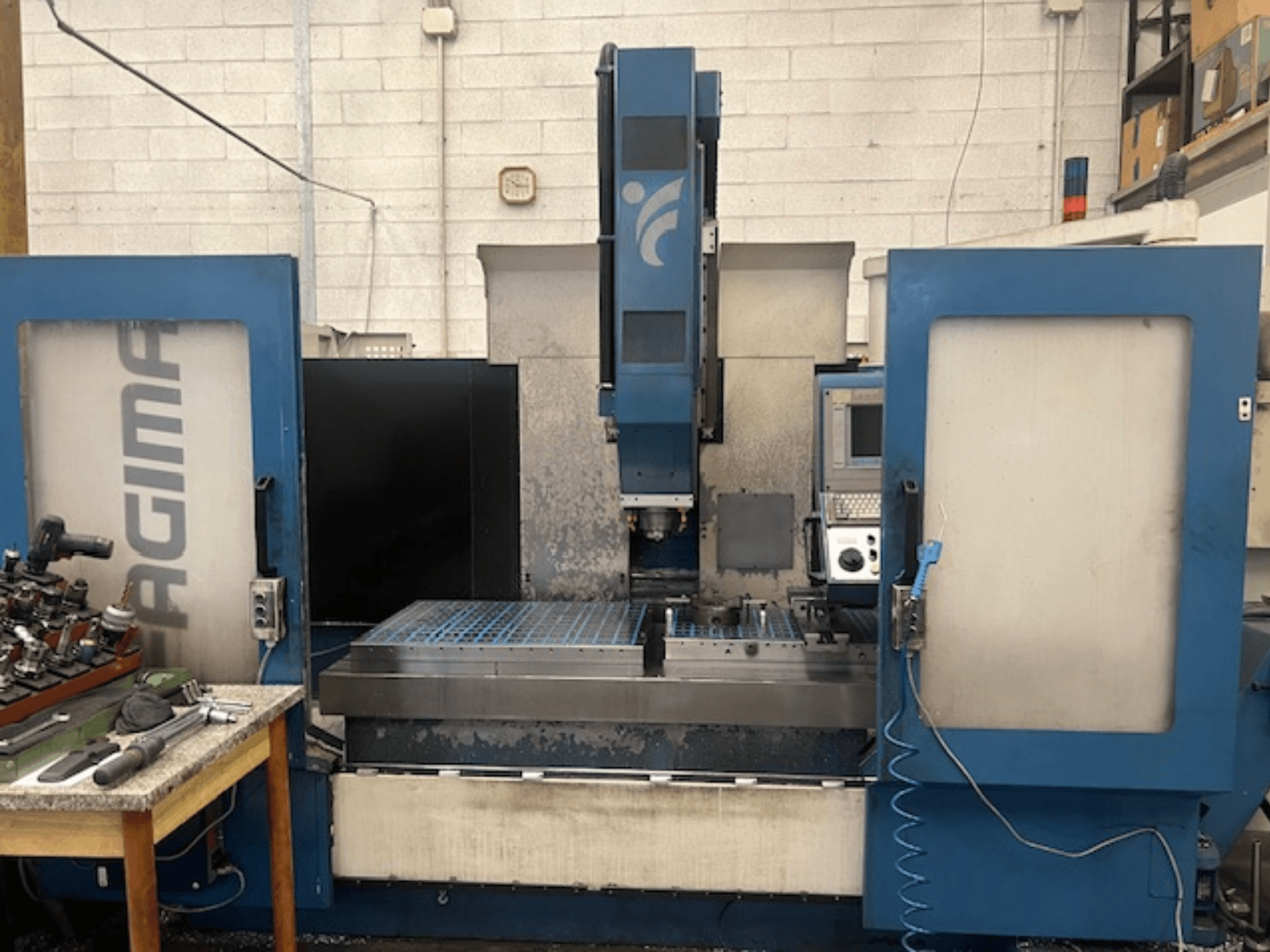 Vista frontal de uma máquina CNC JAGIMA com uma grande área de trabalho em metal, painel de controlo visível e ferramentas numa mesa próxima.