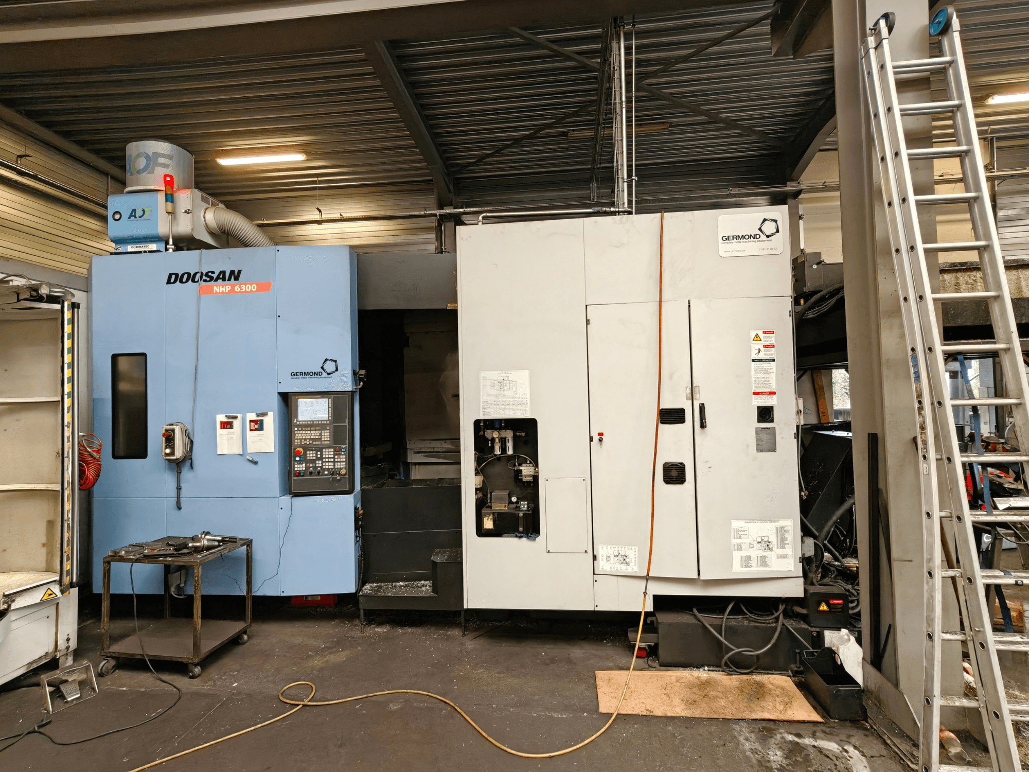 Centro de maquinagem horizontal DOOSAN NHP6300, visto de frente, mostrando o painel de controlo e as máquinas adjacentes num ambiente de oficina.