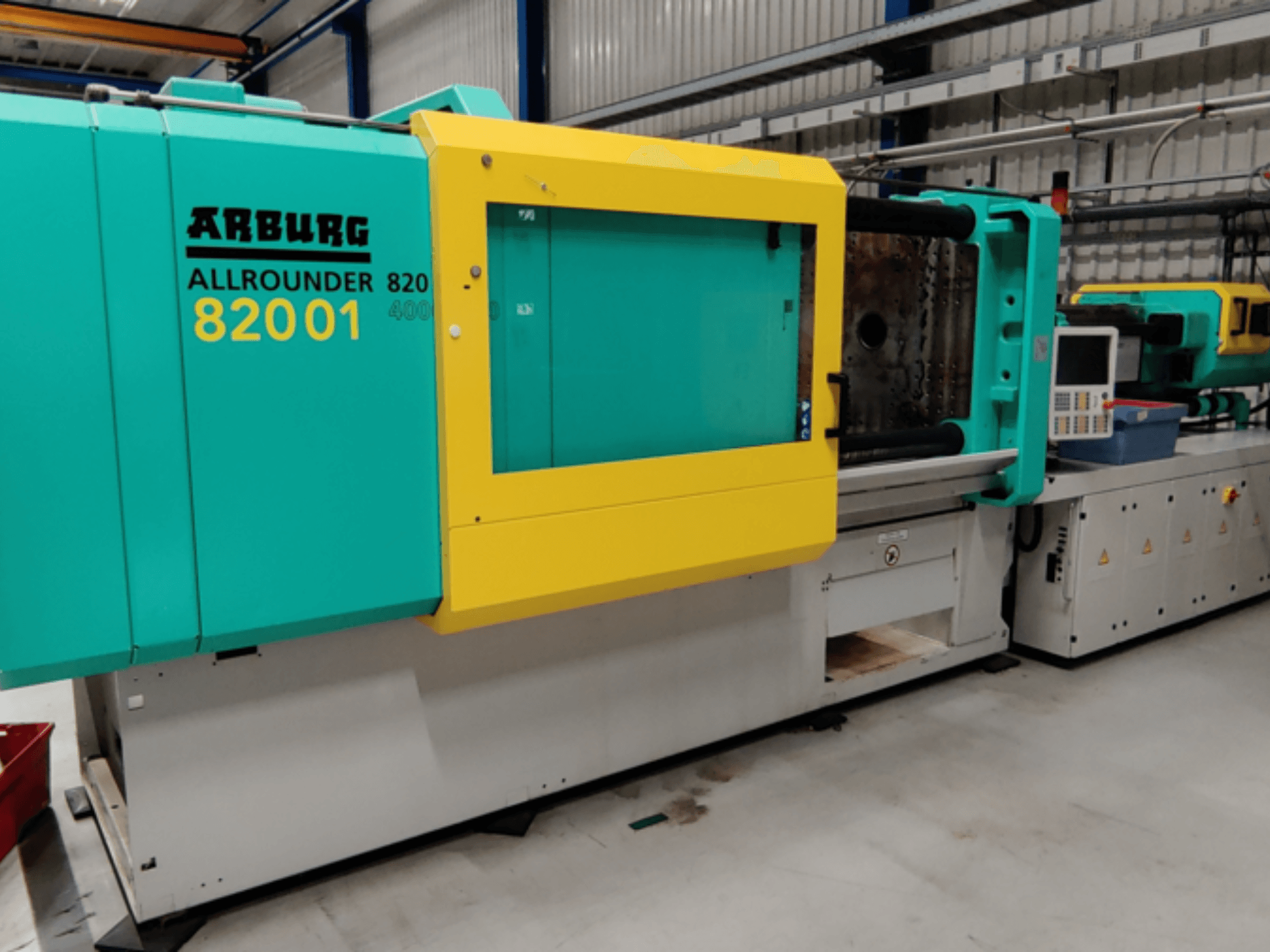 Máquina de moldagem por injeção Arburg ALLROUNDER 82001 em azul-petróleo e amarelo, vista lateral mostrando o painel de comando e as caraterísticas operacionais.
