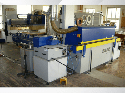 Máquina de colar bordas FELDER G500 numa oficina, vista lateral que mostra a estrutura azul e amarela com os comandos e a rede de condutas.