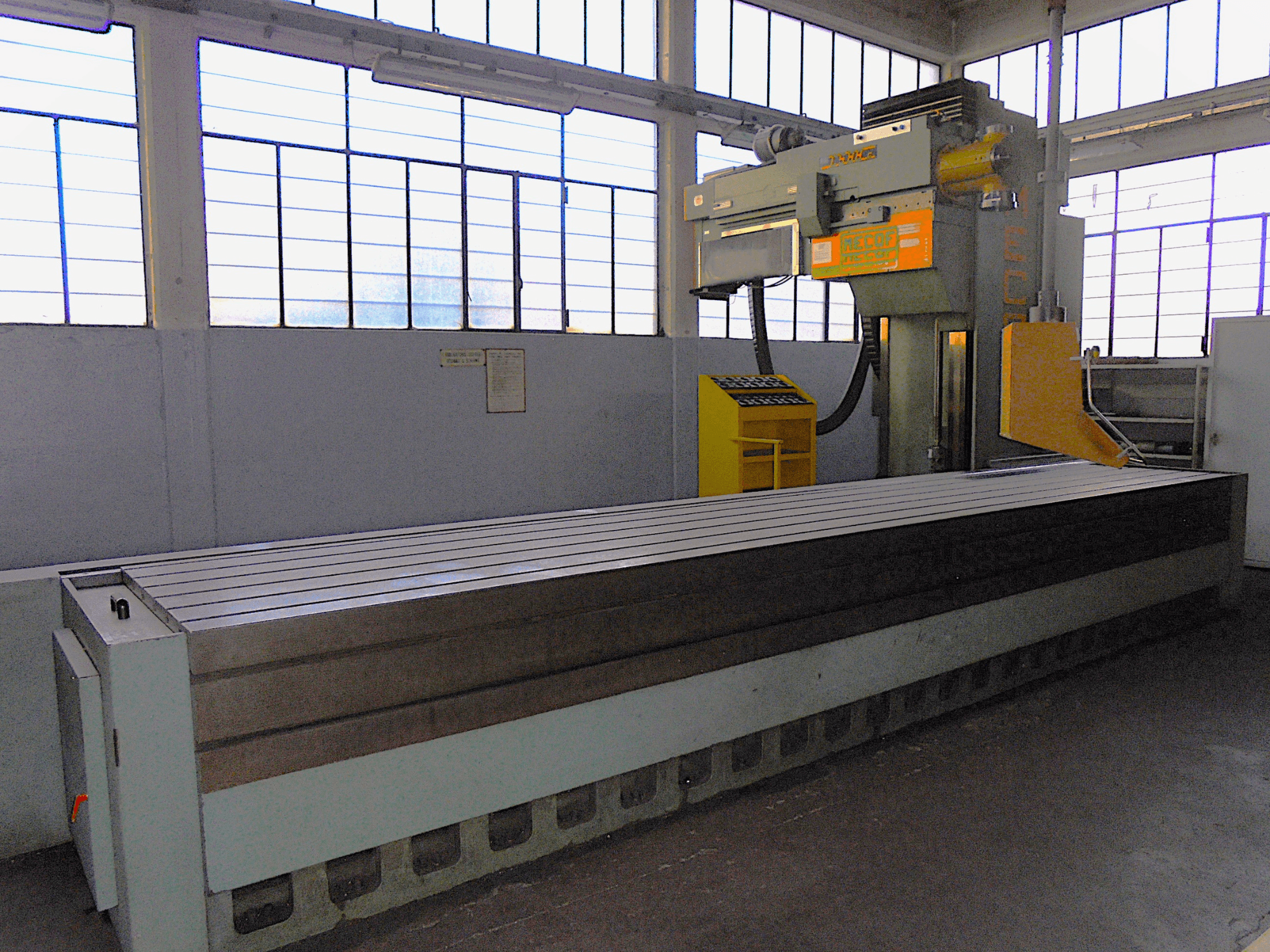 Fresadora vertical de marca desconhecida numa fábrica, vista lateral, apresenta uma mesa grande com superfície ranhurada e painel de controlo.