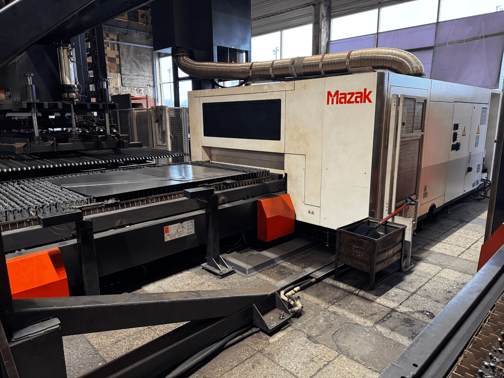 Máquina de corte a laser Mazak numa oficina, vista lateral mostrando o painel de controlo, o transportador e um recipiente de recolha.