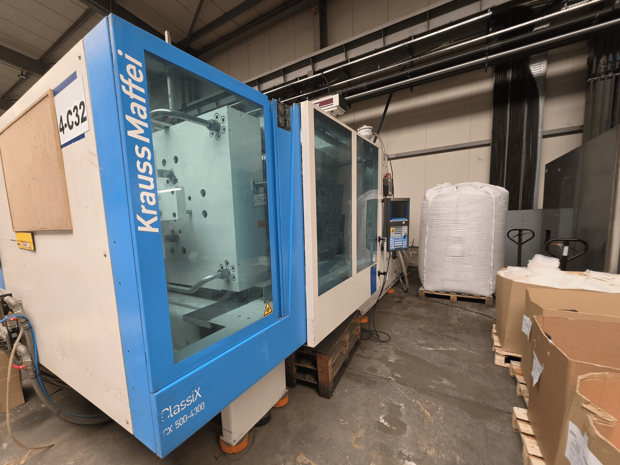 Máquina de moldagem por injeção hidráulica Krauss Maffei 500-4000 CX, vista lateral, mostrando o painel de controlo e os materiais de embalagem próximos.