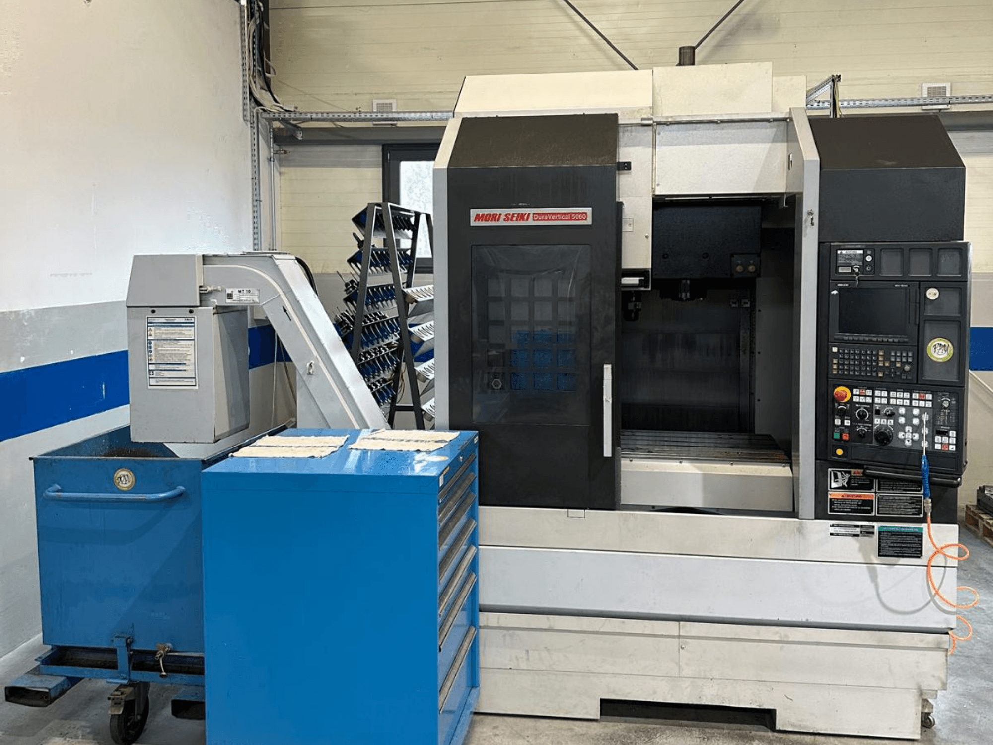 MORI SEIKI DURA VERTICAL 5060, vista frontal de um centro de maquinagem vertical com painel de controlo e armazém de ferramentas nas proximidades.