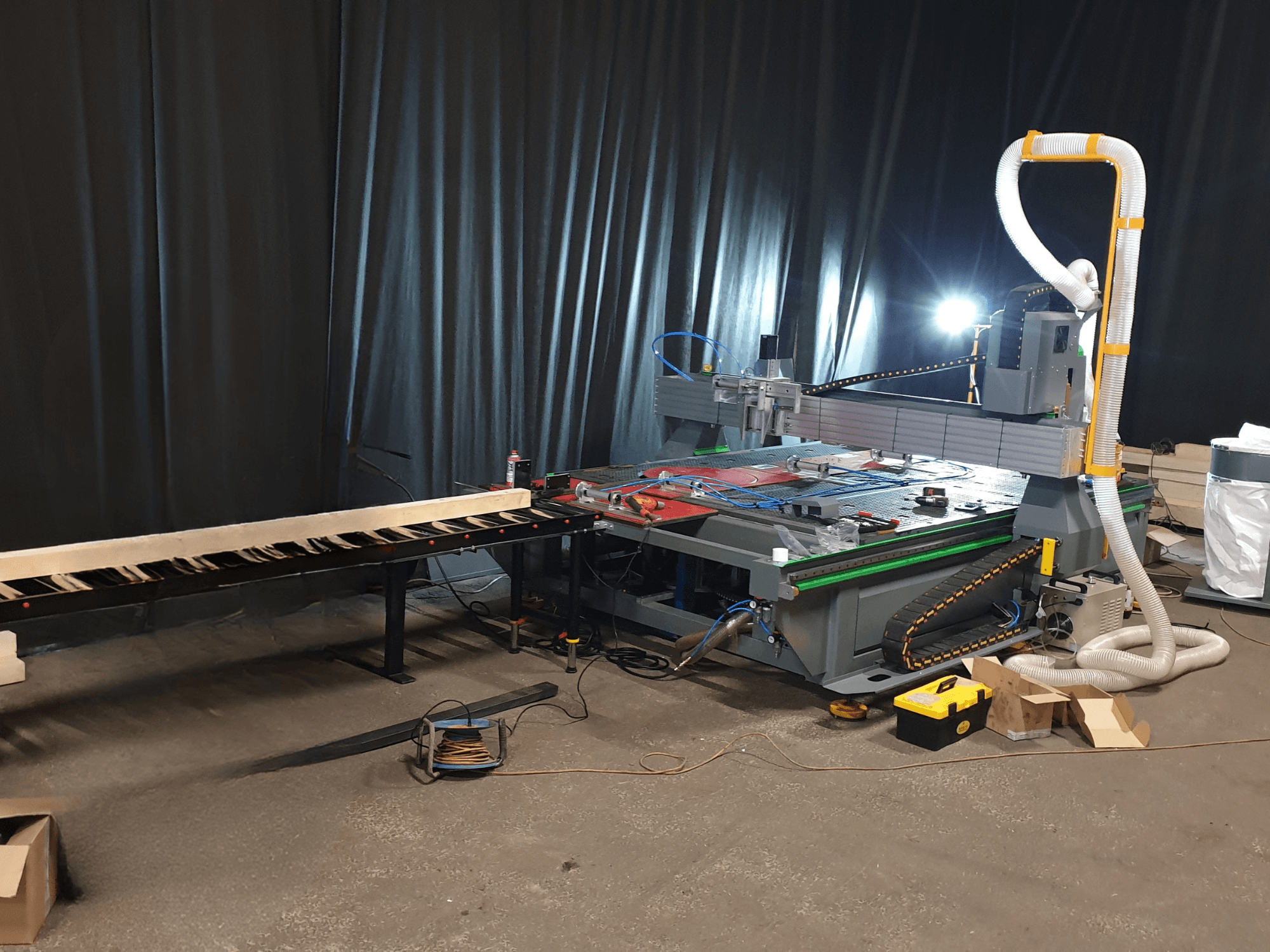 Máquina CNC com um transportador lateral, ferramentas na mesa e conduta para recolha de poeiras. Fundo preto, vista frontal.