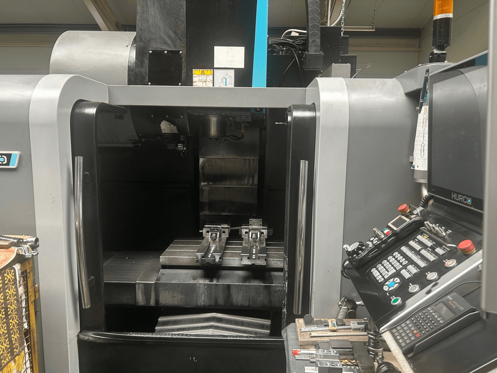 Vista frontal do centro de maquinação CNC Hurco, mostrando o suporte da ferramenta, a peça de trabalho em metal e o painel de controlo com botões e visor.