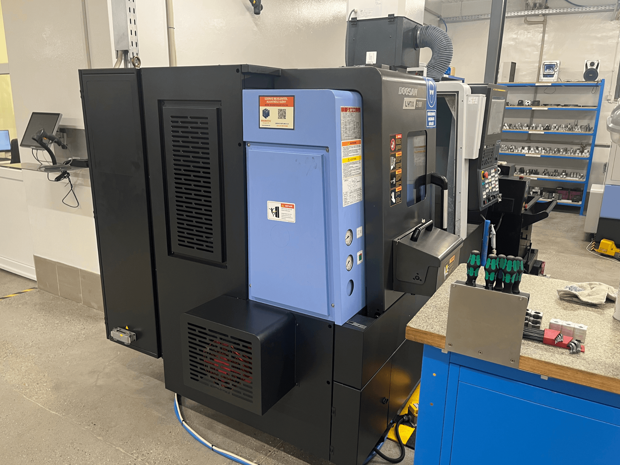 Torno Doosan Lynx 2100, vista lateral, mostrando o painel de controlo, o sistema de ventilação e as ferramentas numa bancada de trabalho.