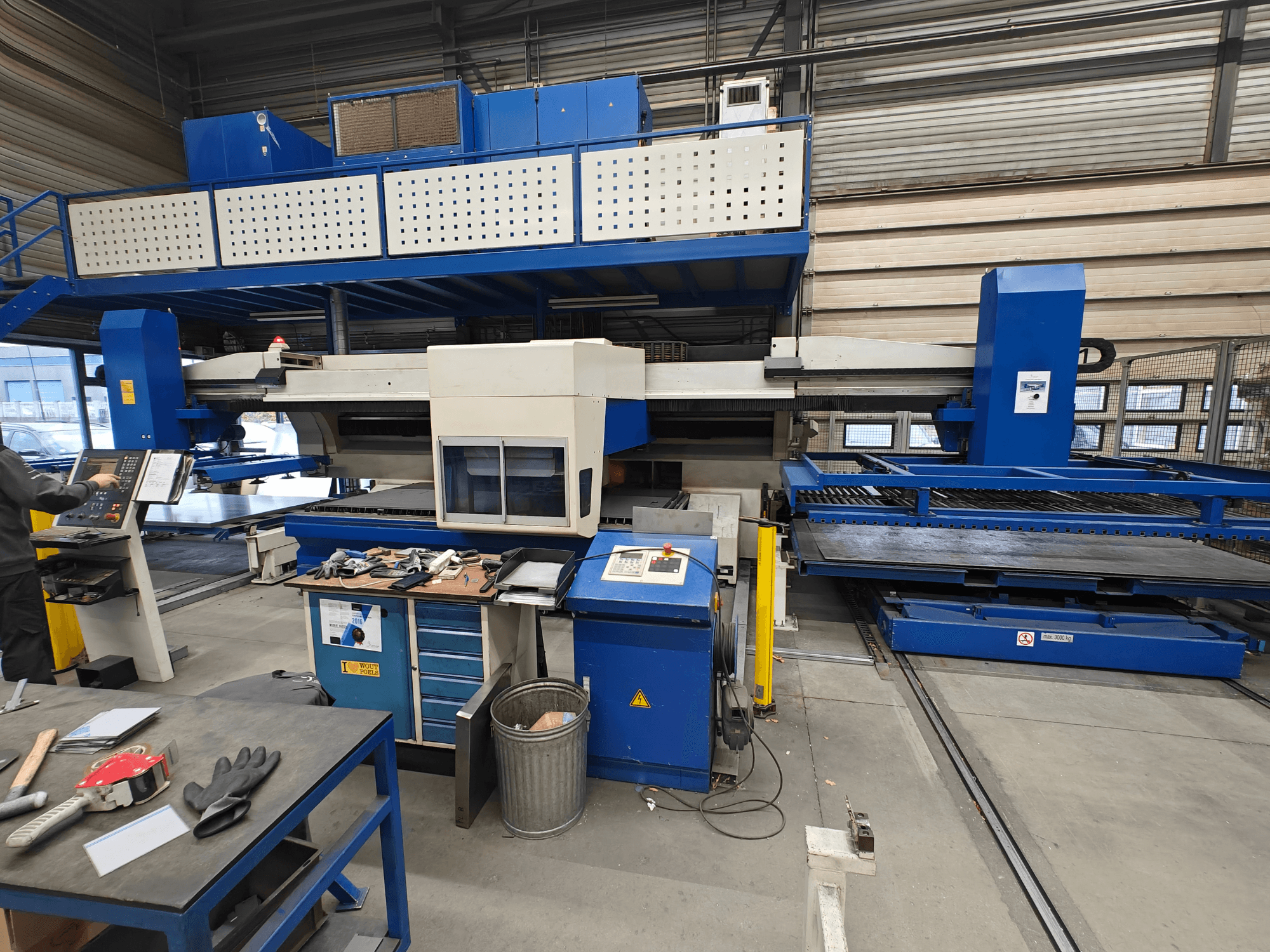 Máquina CNC industrial vista de frente, com construção em azul e branco, painel de controlo e várias ferramentas na mesa de trabalho.