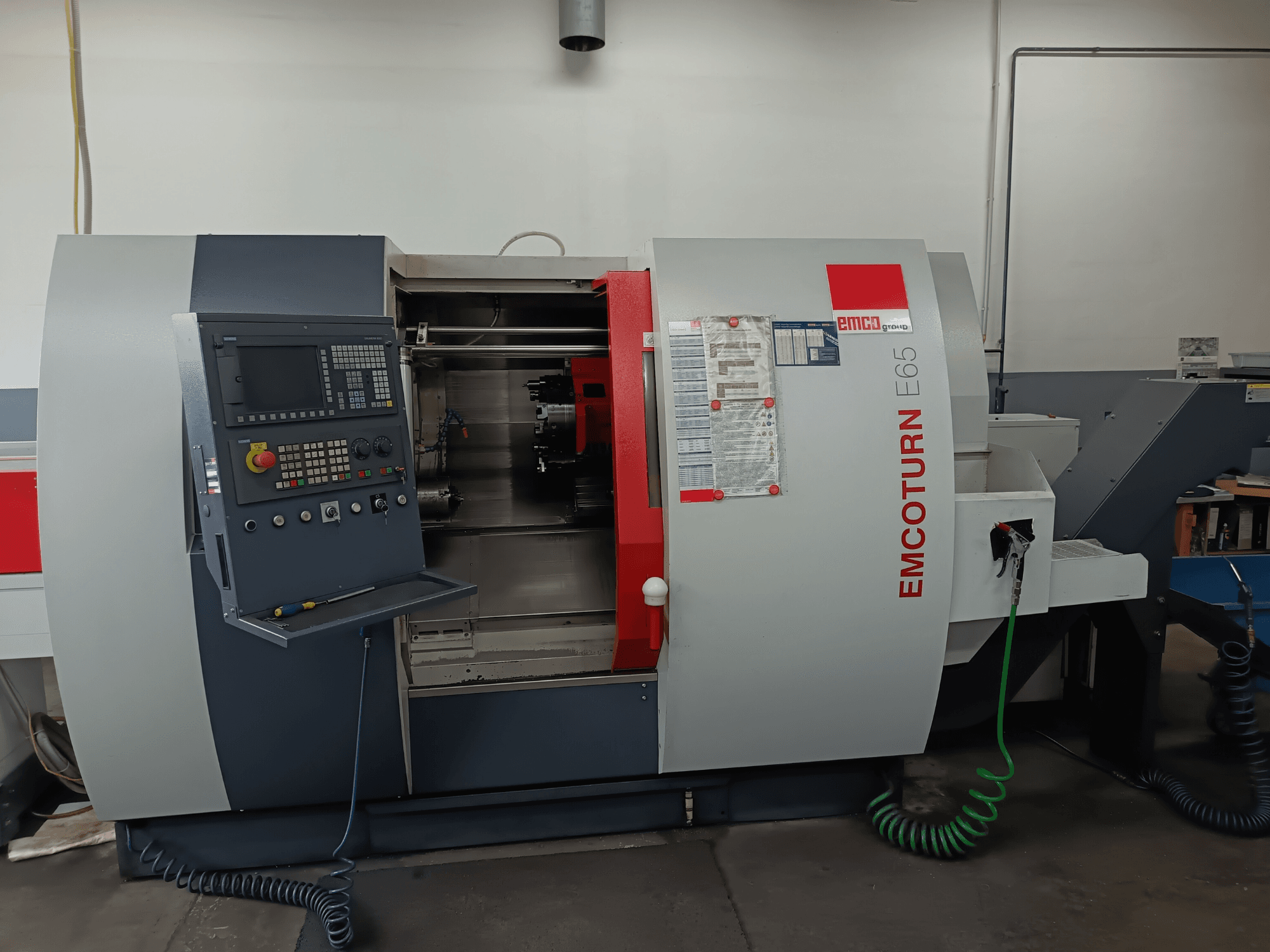 Centro de torneamento EMCO EMCOTURN E65, vista frontal mostrando o painel de controlo, mecanismo interno e caraterísticas numa oficina.
