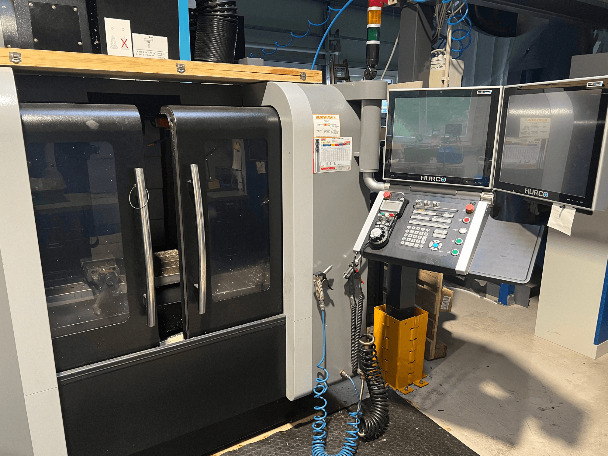 Vista frontal da máquina CNC Hurco com dois monitores e painel de controlo. Possui portas transparentes e área de maquinação visível no interior.