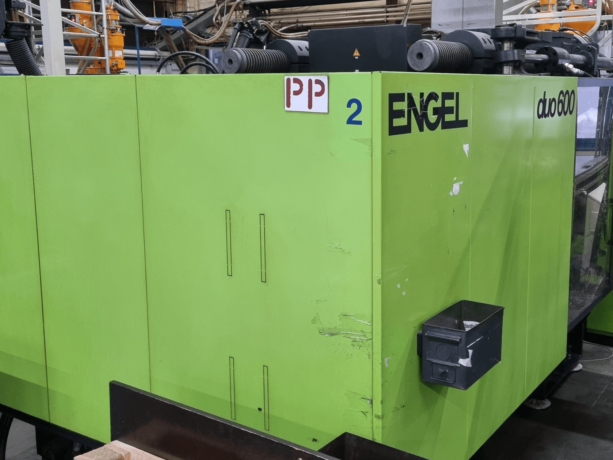 Máquina de moldagem por injeção hidráulica Engel Duo 600, exterior verde, vista frontal, com peças identificadas e caixa de armazenamento de material.