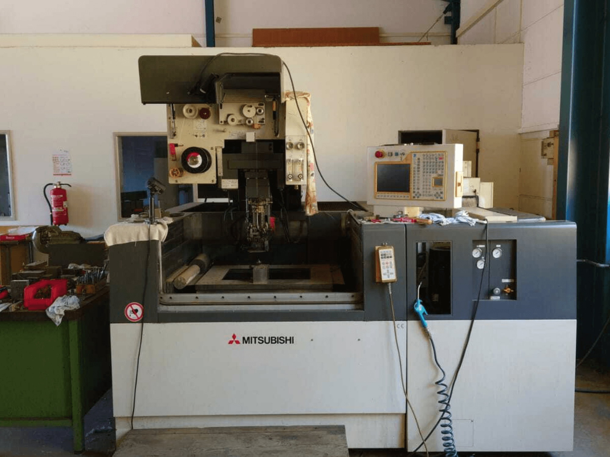 Vista frontal da máquina de eletroerosão por fio Mitsubishi FA20, com o painel de controlo, a área de trabalho e a sinalização de segurança visíveis.