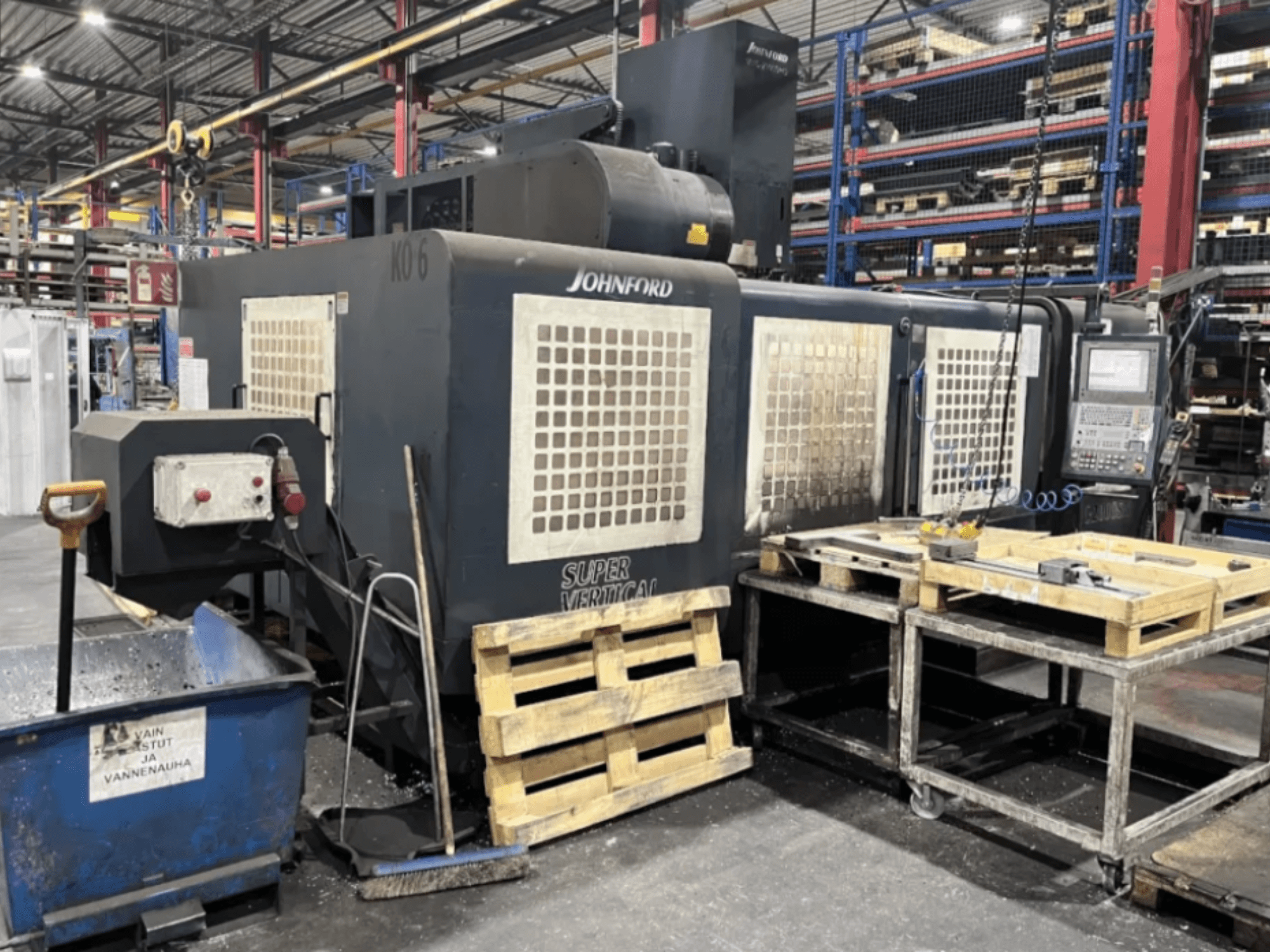 Fresadora Super Vertical Johnford numa fábrica, vista lateral com painel de controlo, paletes e equipamento de limpeza nas proximidades.