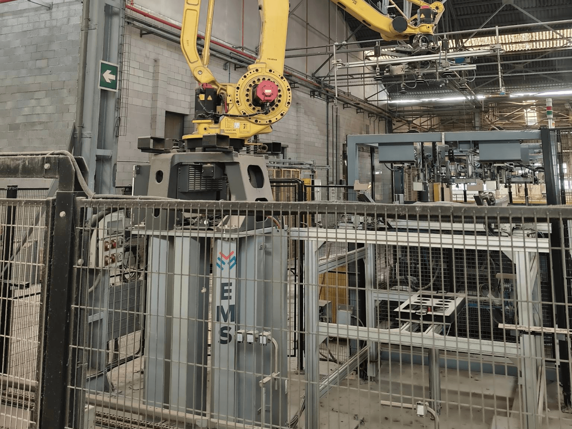 Vista frontal do braço robótico FANUC M-410iC/315 posicionado sobre uma ferramenta EMS, com equipamento industrial e uma vedação ao fundo.