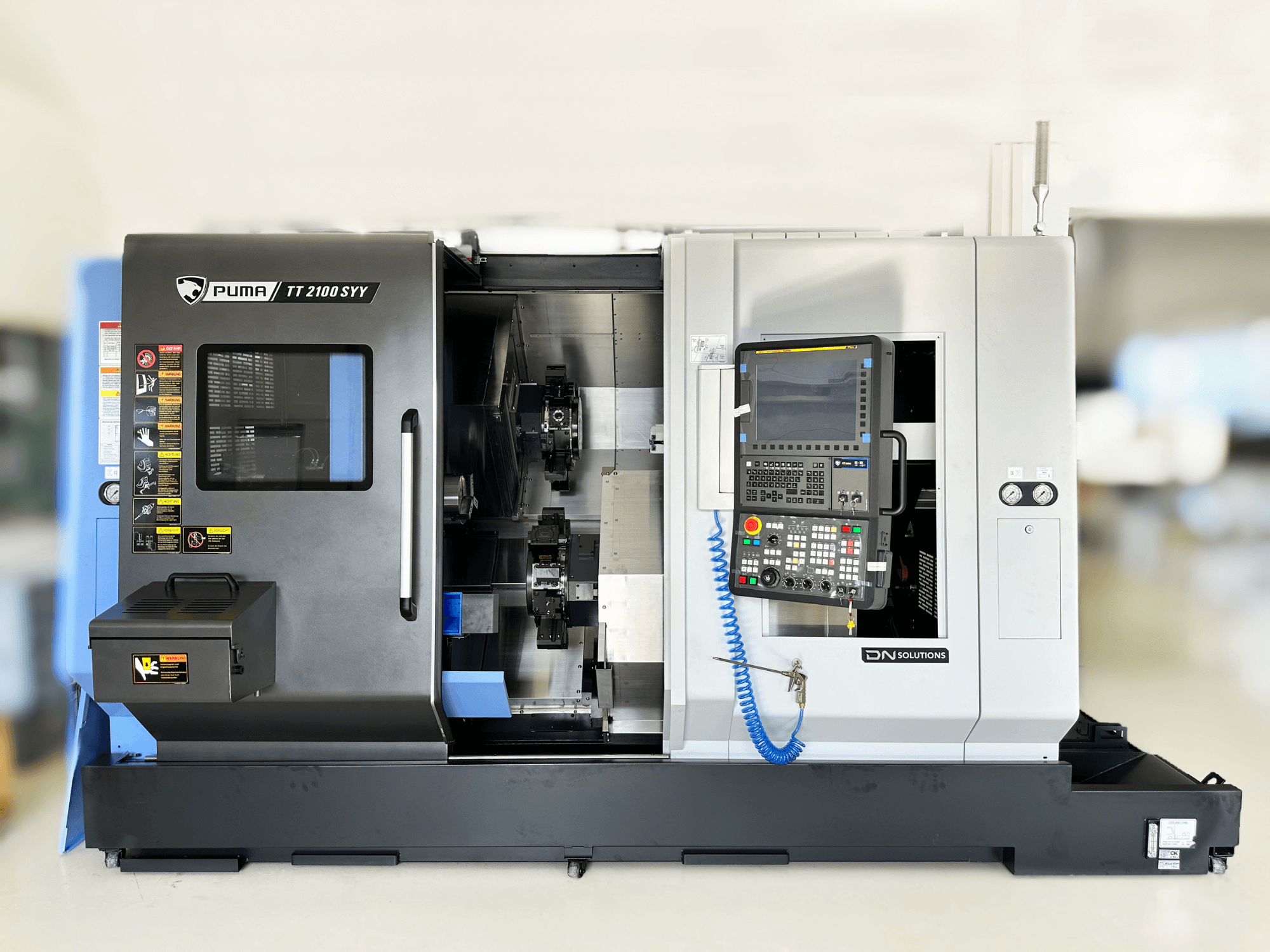 Centro de torneamento-fresagem DN SOLUTIONS PUMA TT2100SYY vista frontal, com um painel de controlo e componentes interiores visíveis.