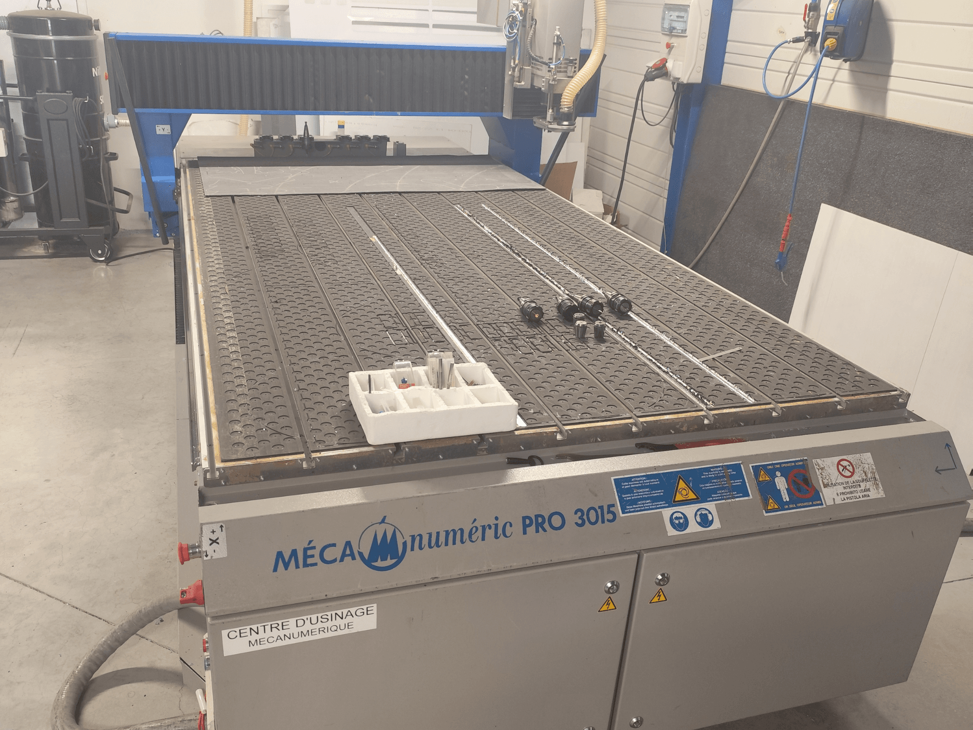Vista frontal da fresadora CNC MECANUMERIC Pro 3015, apresentando uma mesa de corte detalhada, o painel de controlo e os acessórios colocados sobre a mesa.