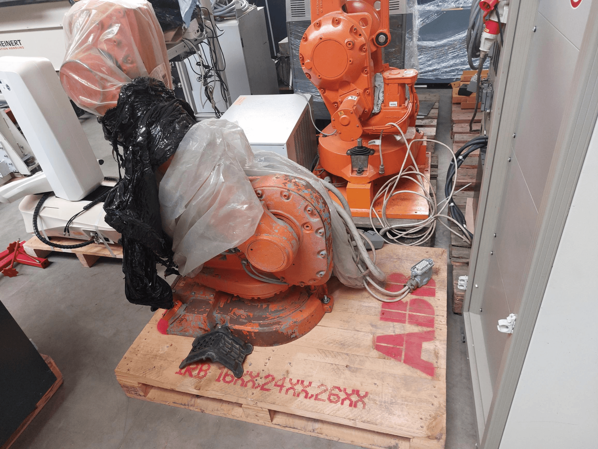 Braço do robot ABB IRB 1600 M2004 parcialmente coberto de plástico, visto de frente, com cabos e maquinaria adicional nas proximidades.
