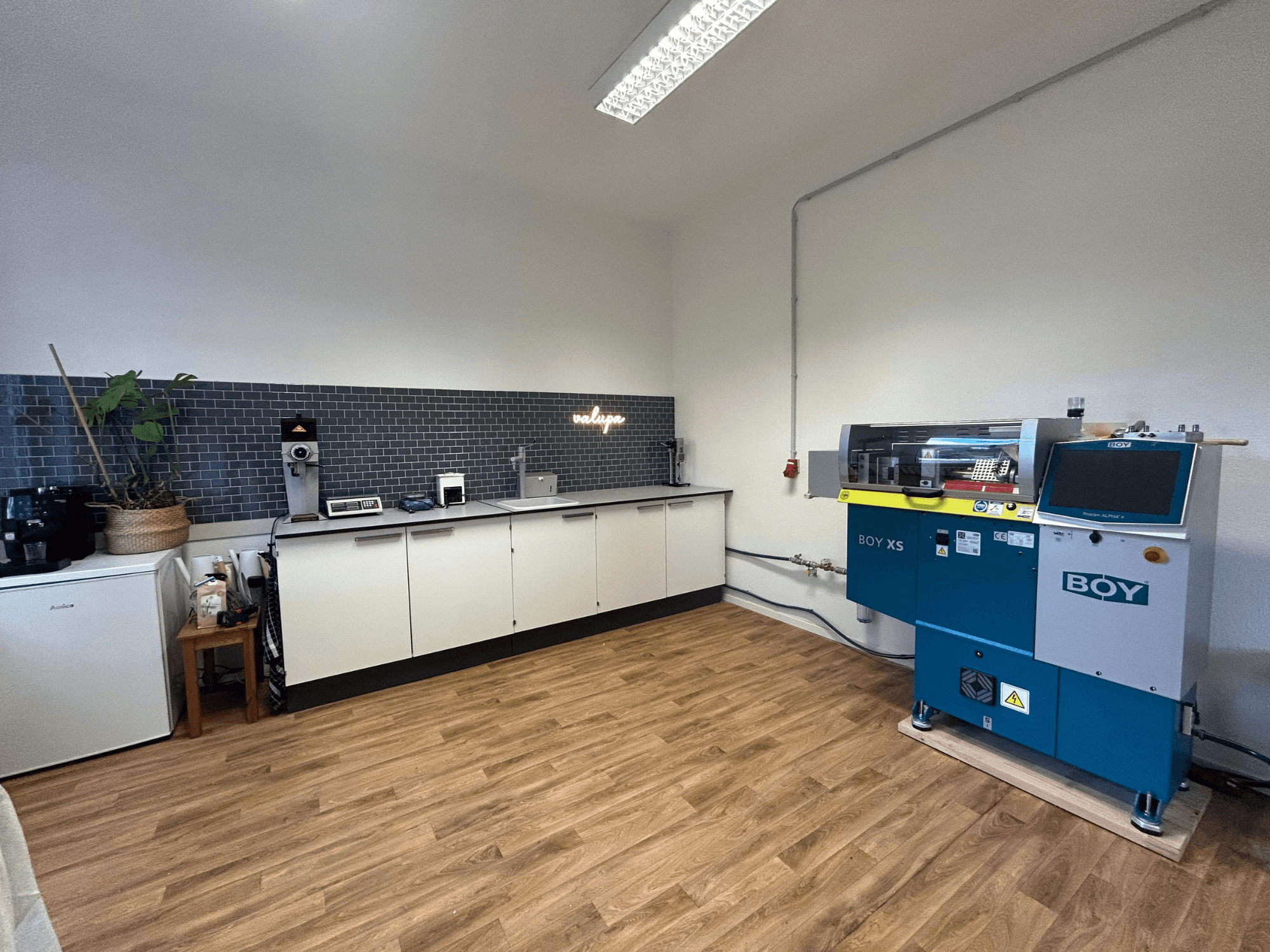 Máquina de moldagem por injeção hidráulica BOY XS 161, vista frontal, numa cozinha moderna com electrodomésticos e chão de madeira.