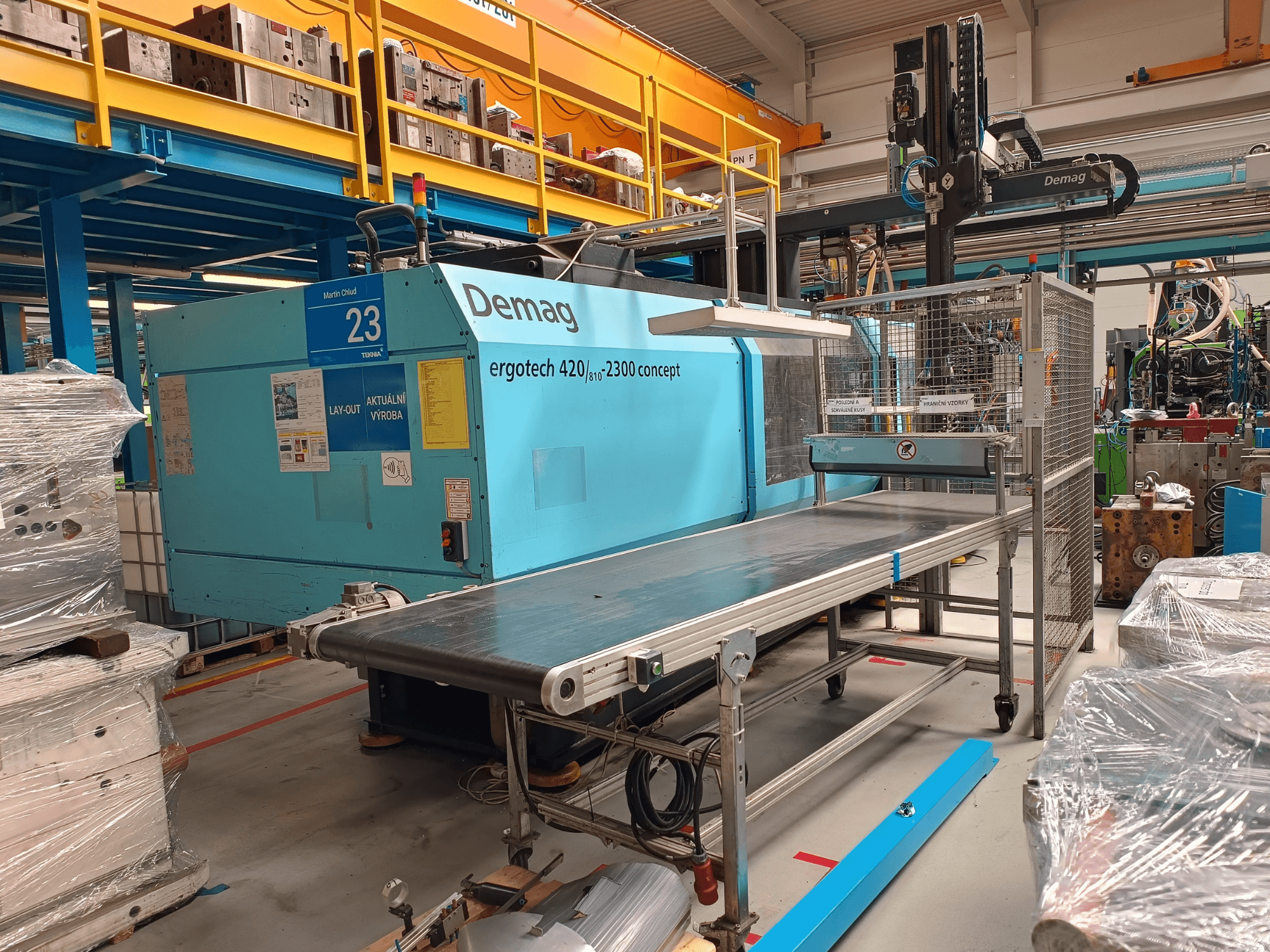 Máquina de moldagem por injeção hidráulica Demag ergotech 420/810 - 2300 em vista lateral, com um transportador e uma caixa de segurança.