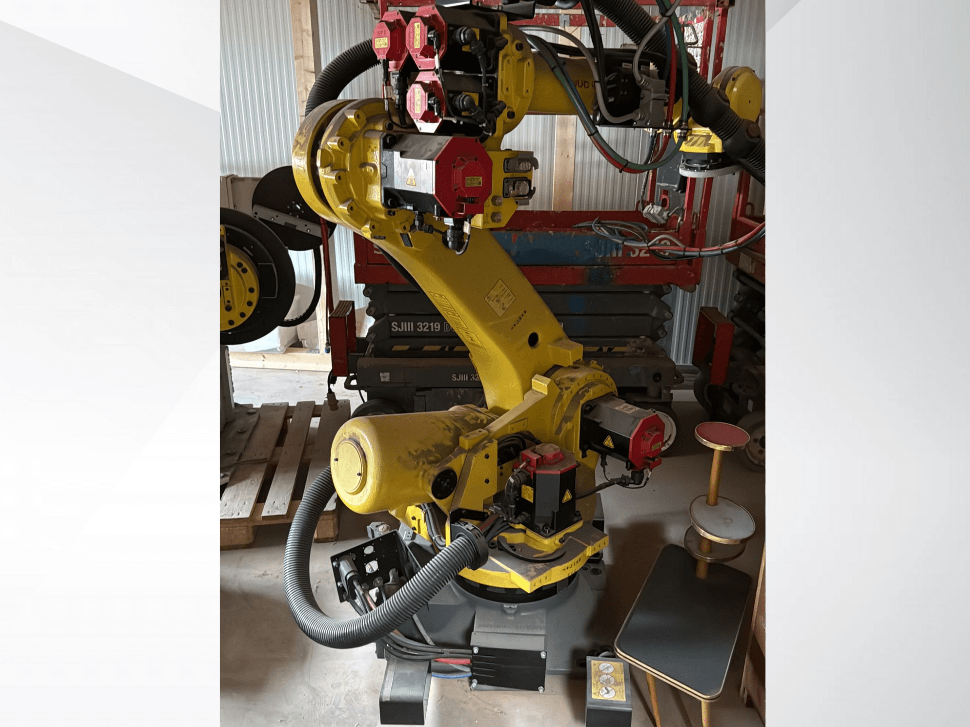 Braço robótico industrial amarelo com controladores vermelhos, vista frontal; possui várias articulações e mangueiras, colocado num local de trabalho.