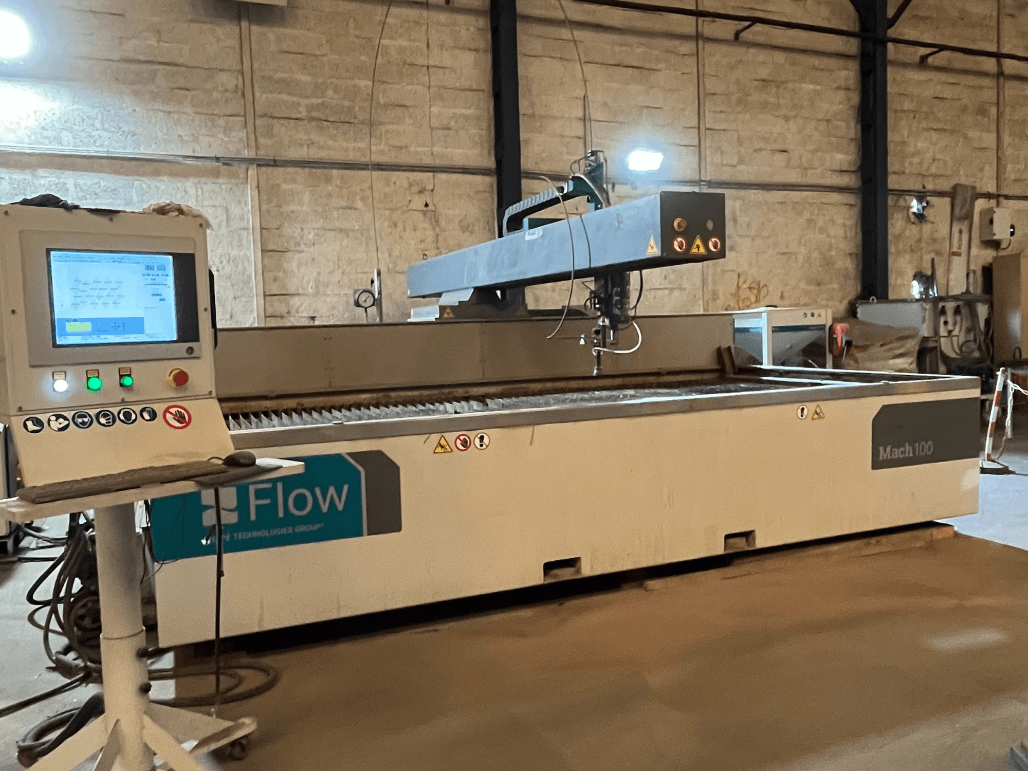 Cortador de jato de água Flow Mach 100, vista lateral, com um painel de controlo digital e uma cabeça de corte visíveis numa mesa de trabalho.