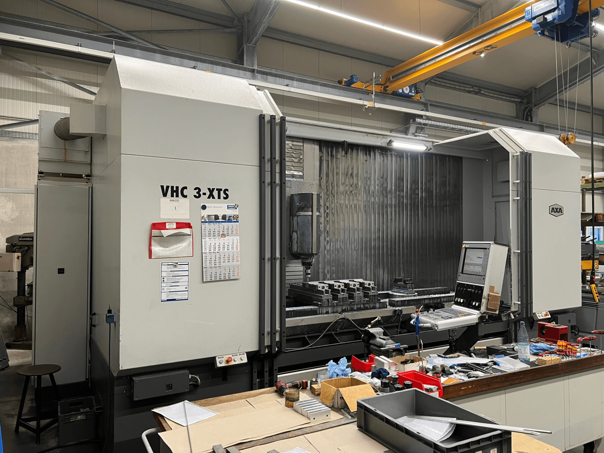 Máquina CNC AXA VHC 3-XTS em vista lateral, com painel de controlo e área de trabalho repleta de ferramentas e componentes.