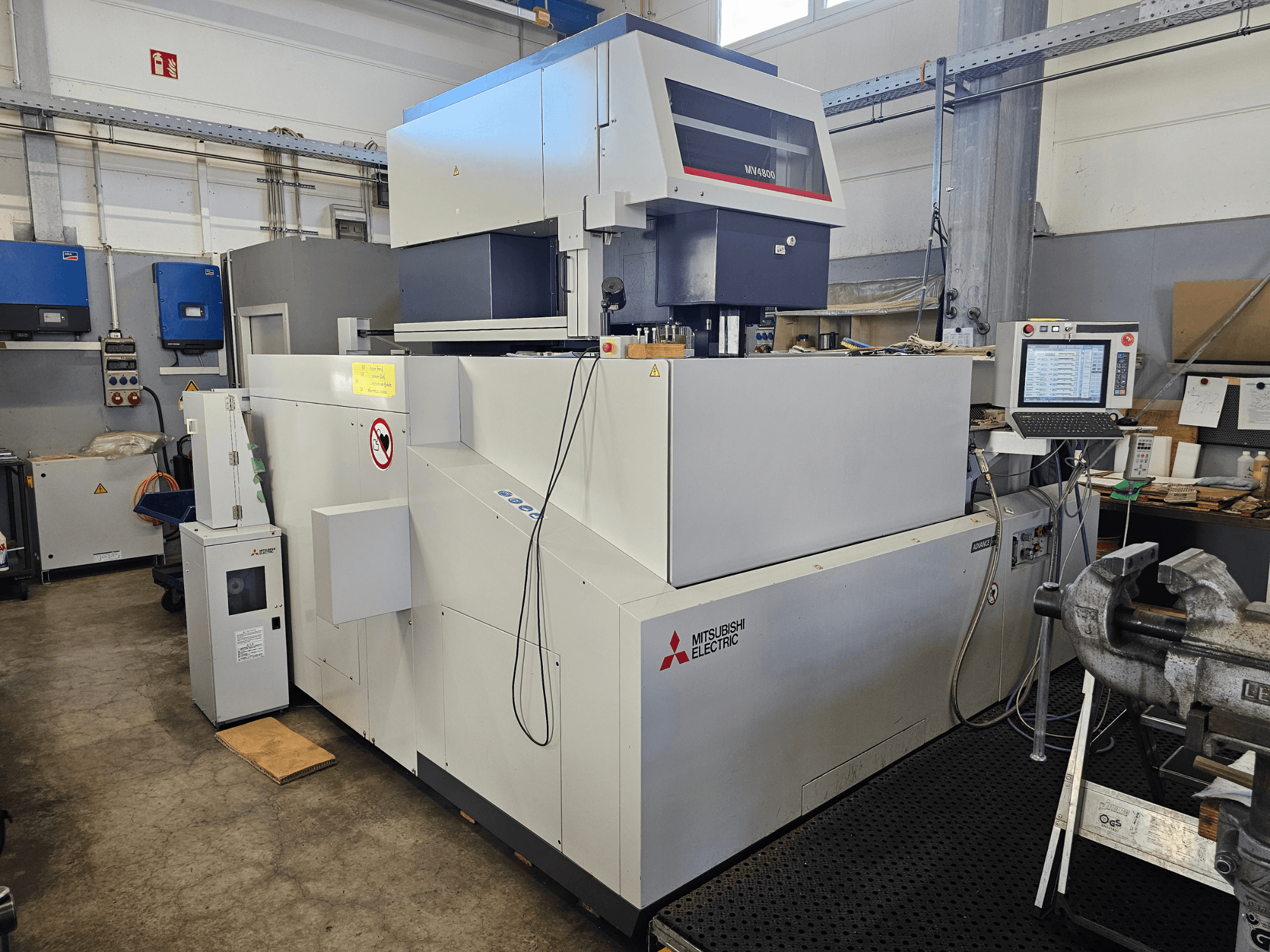 Máquina EDM de fio Mitsubishi MV4800 numa oficina, vista lateral mostrando o painel de controlo, etiquetas de segurança e equipamento circundante.