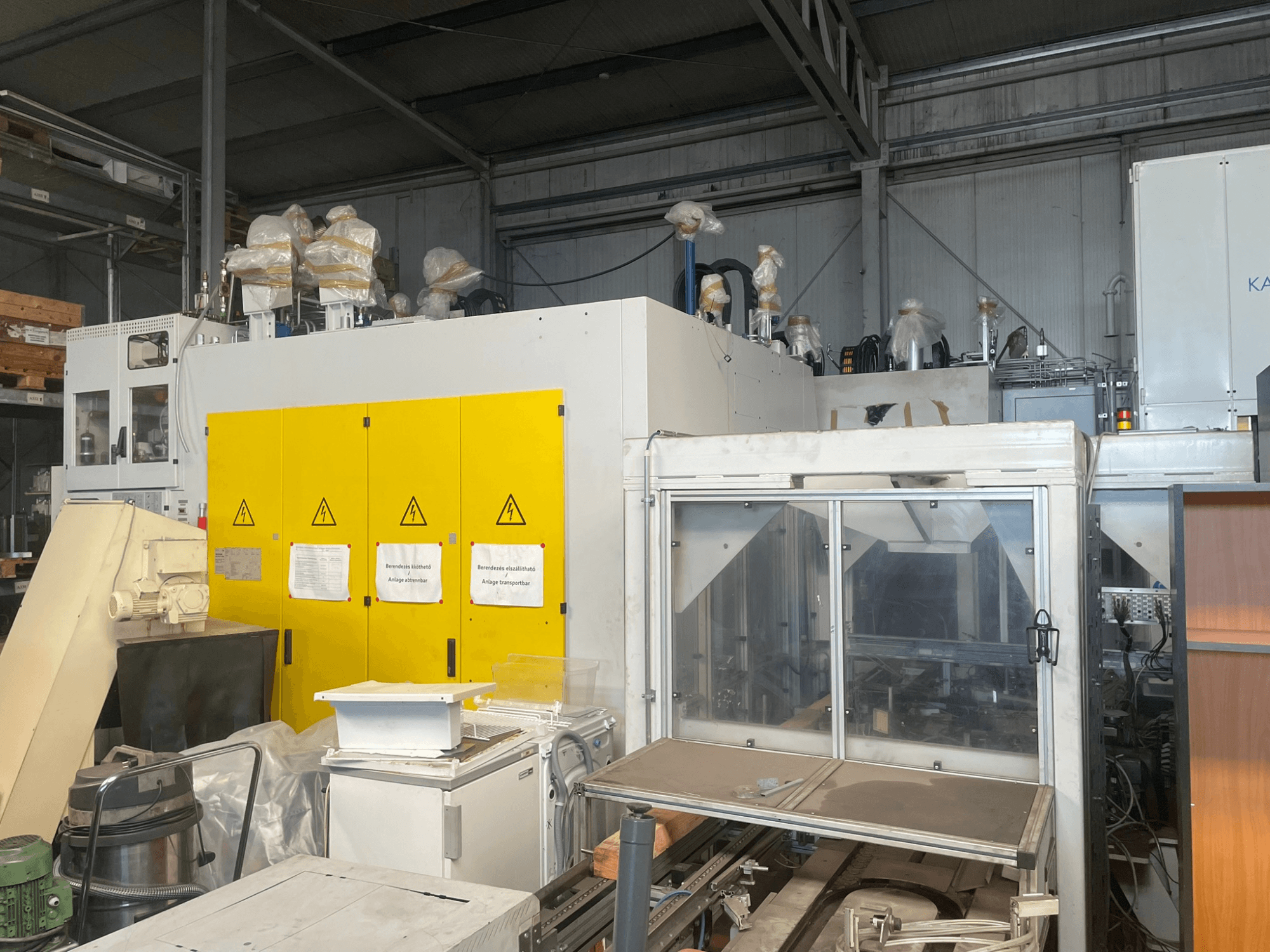 Máquinas industriais com painel de controlo amarelo, vista lateral, com etiquetas de aviso e revestimentos de plástico, num armazém.